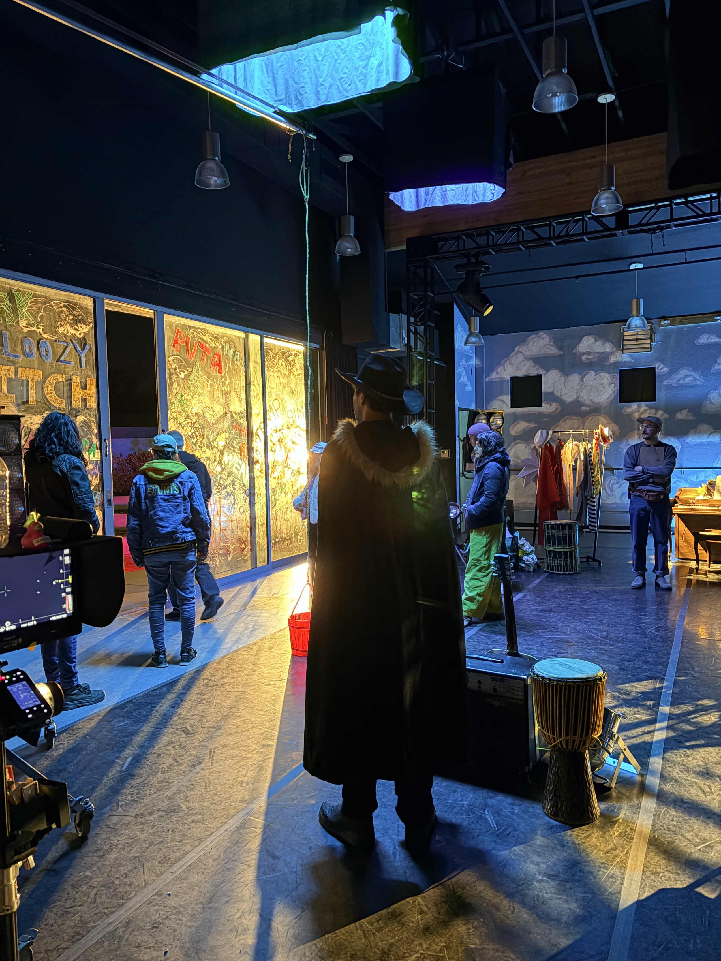 People gathering inside a dark, artistic space with vibrant lighting, wall graffiti, a clothing rack, and musical instruments including a drum. Some individuals are engaged in photography or filming.