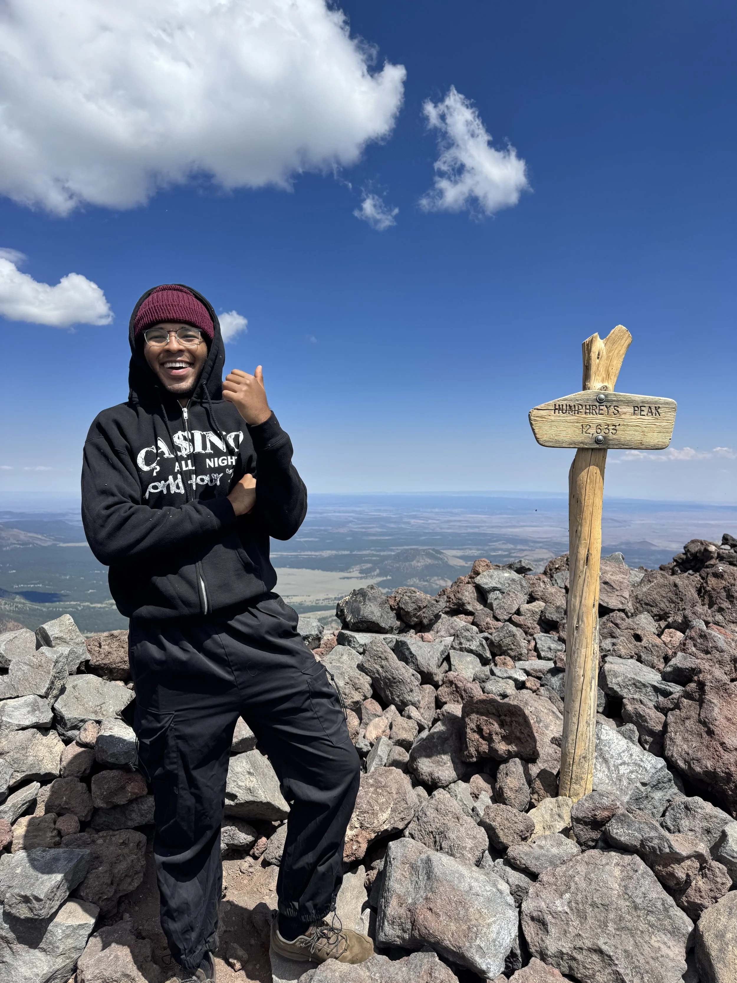 Person smiling and pointing, standing on rocky ground at the summit of Humphreys Peak, with a clear blue sky and clouds in the background, and a wooden sign indicating the peak's name and elevation.