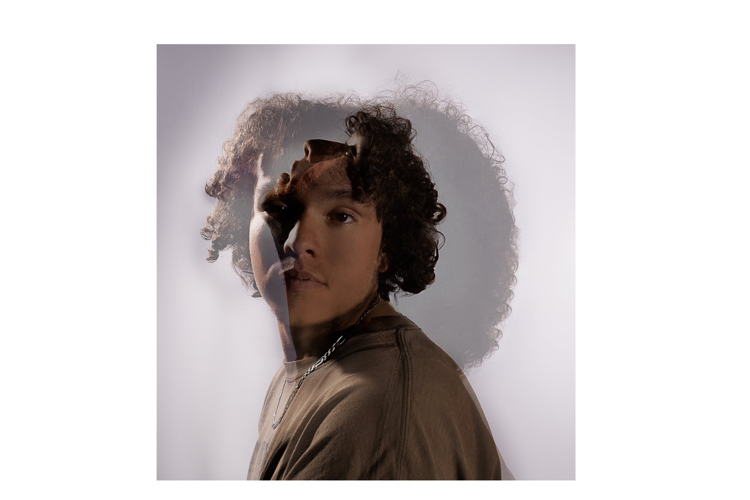 A young person with curly brown hair wearing a brown shirt, silver chain, and necklace, looking at the camera with a neutral expression, against a gray background with a light shadow.