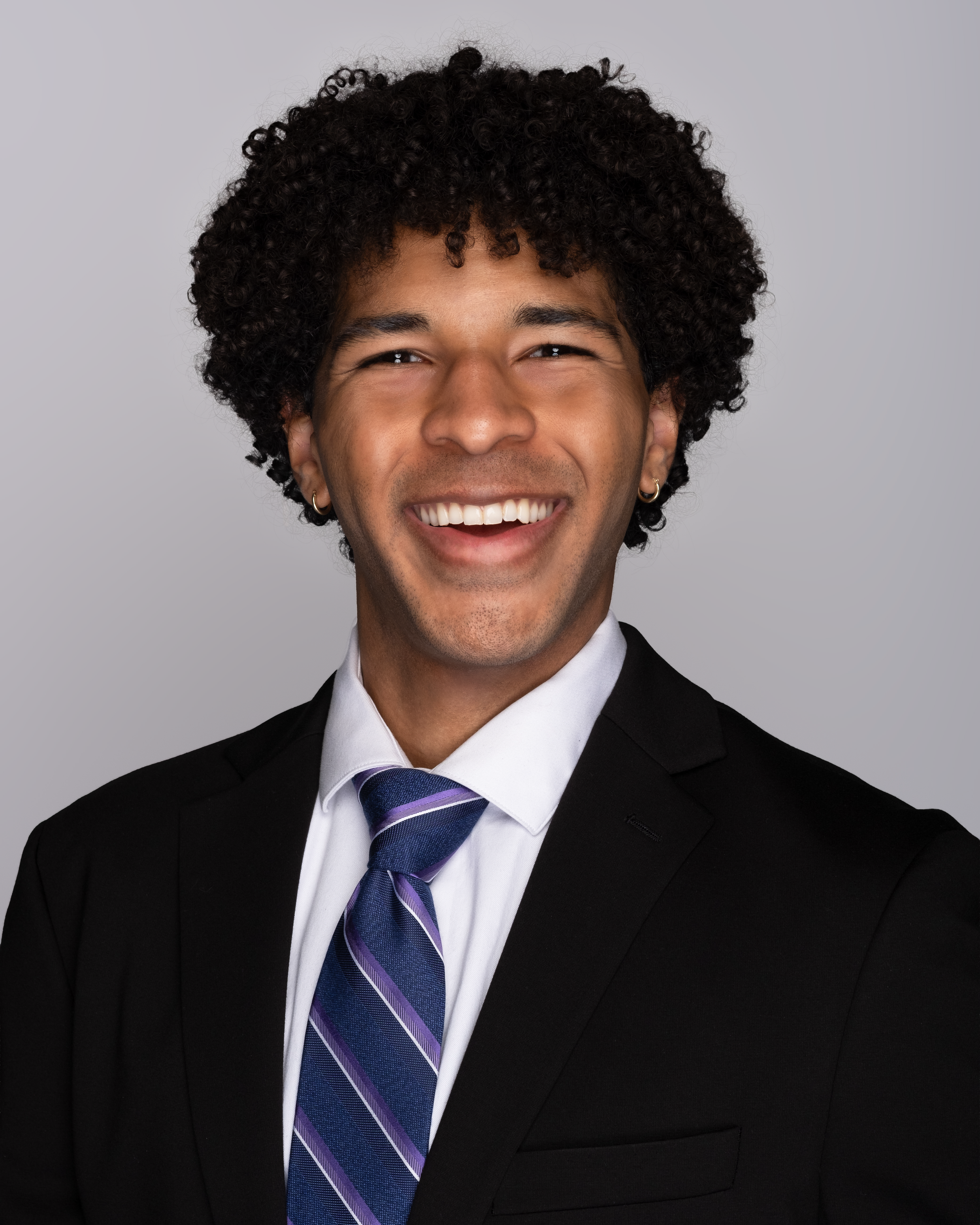 Smiling man wearing a business suit, white shirt, and tie, with curly black hair and earrings, against a gray background.