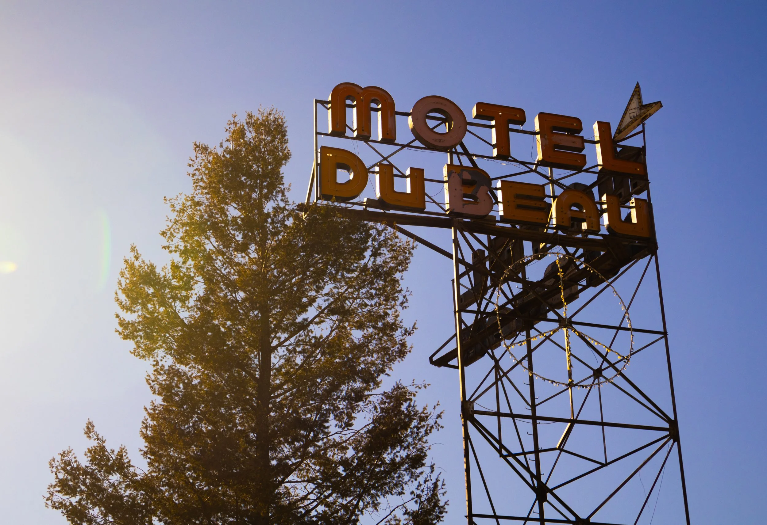 An old motel sign with the words "Motel" and "Beer" in large yellow letters on a metal structure, with a clear blue sky and a tree in the background.