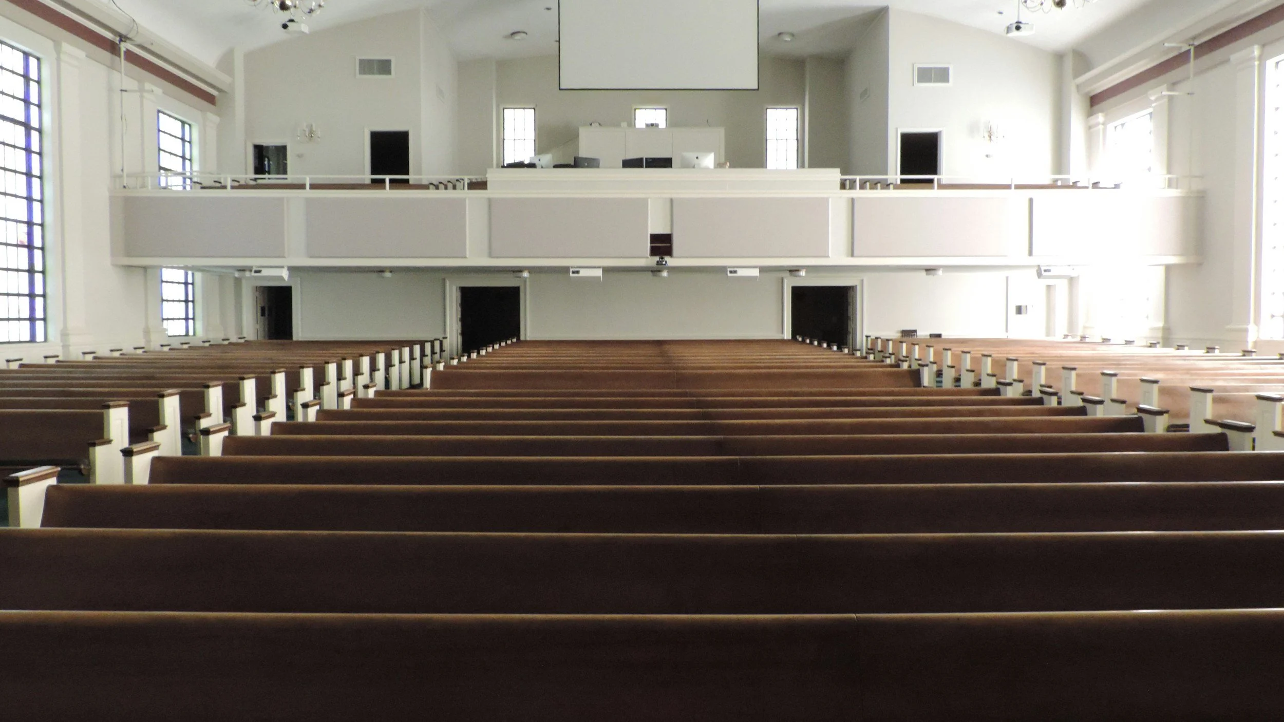 Interior of an empty church with wooden pews, white walls, large windows, and a balcony at the front with a large projection screen hanging from the ceiling.