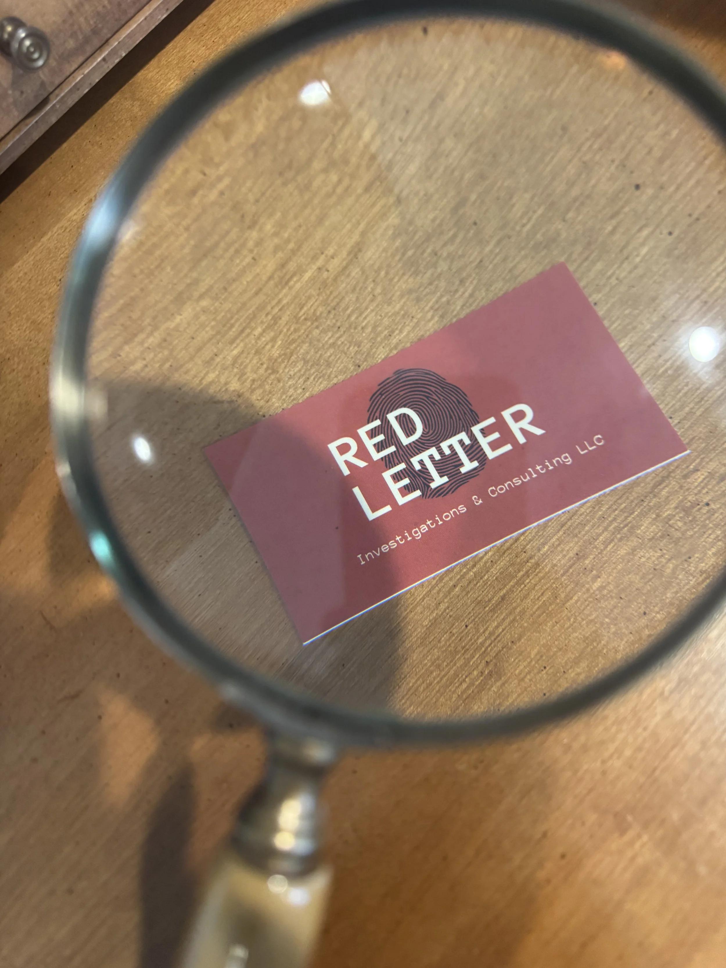Business card for Red Finger Investigations & Consulting LLC viewed through a magnifying glass on wooden surface.