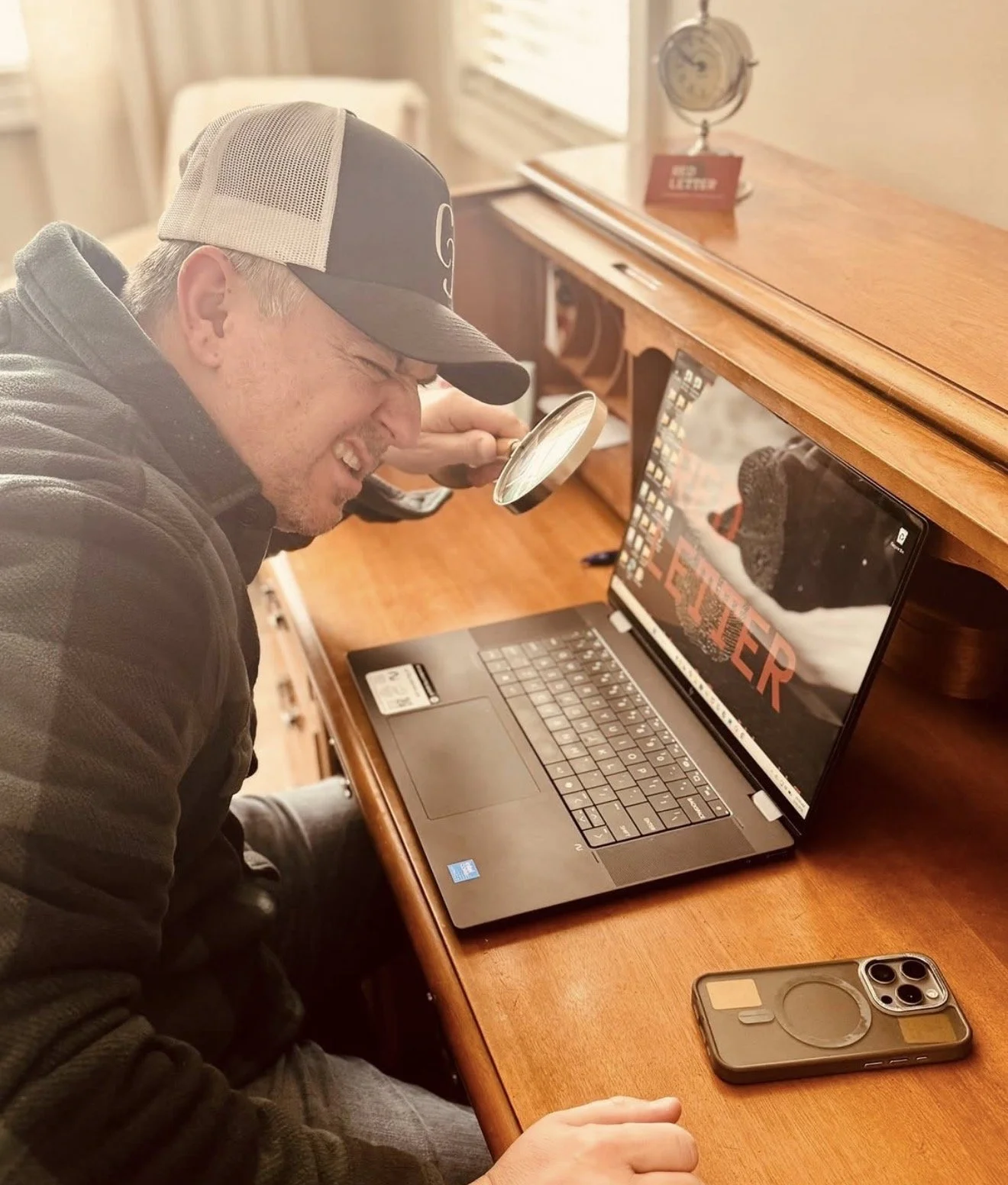 A man with a baseball cap and dark jacket is sitting at a wooden desk, examining a computer screen with a magnifying glass. The computer shows the word "ELEPHANT" with an image of an elephant. On the desk, there is a smartphone and a remote control. The background includes a window with blinds and a clock on the desk.