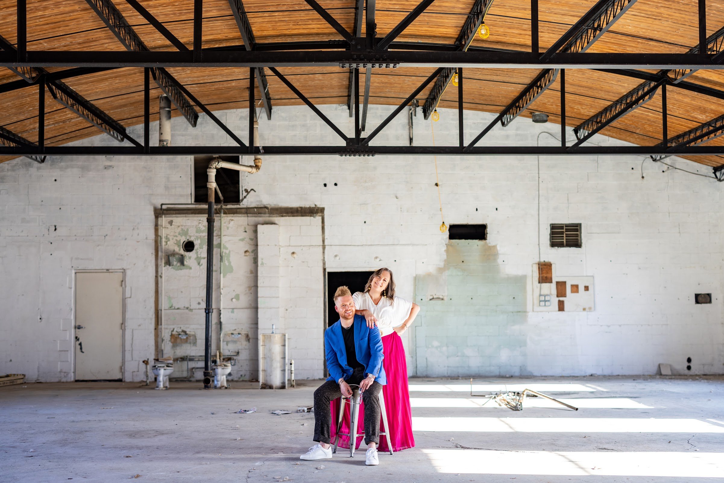 Two people posing in an unfinished industrial space. One person is sitting on a stool, and the other person is standing behind, both smiling. The setting has exposed pipes, unfinished walls, and a wooden ceiling.