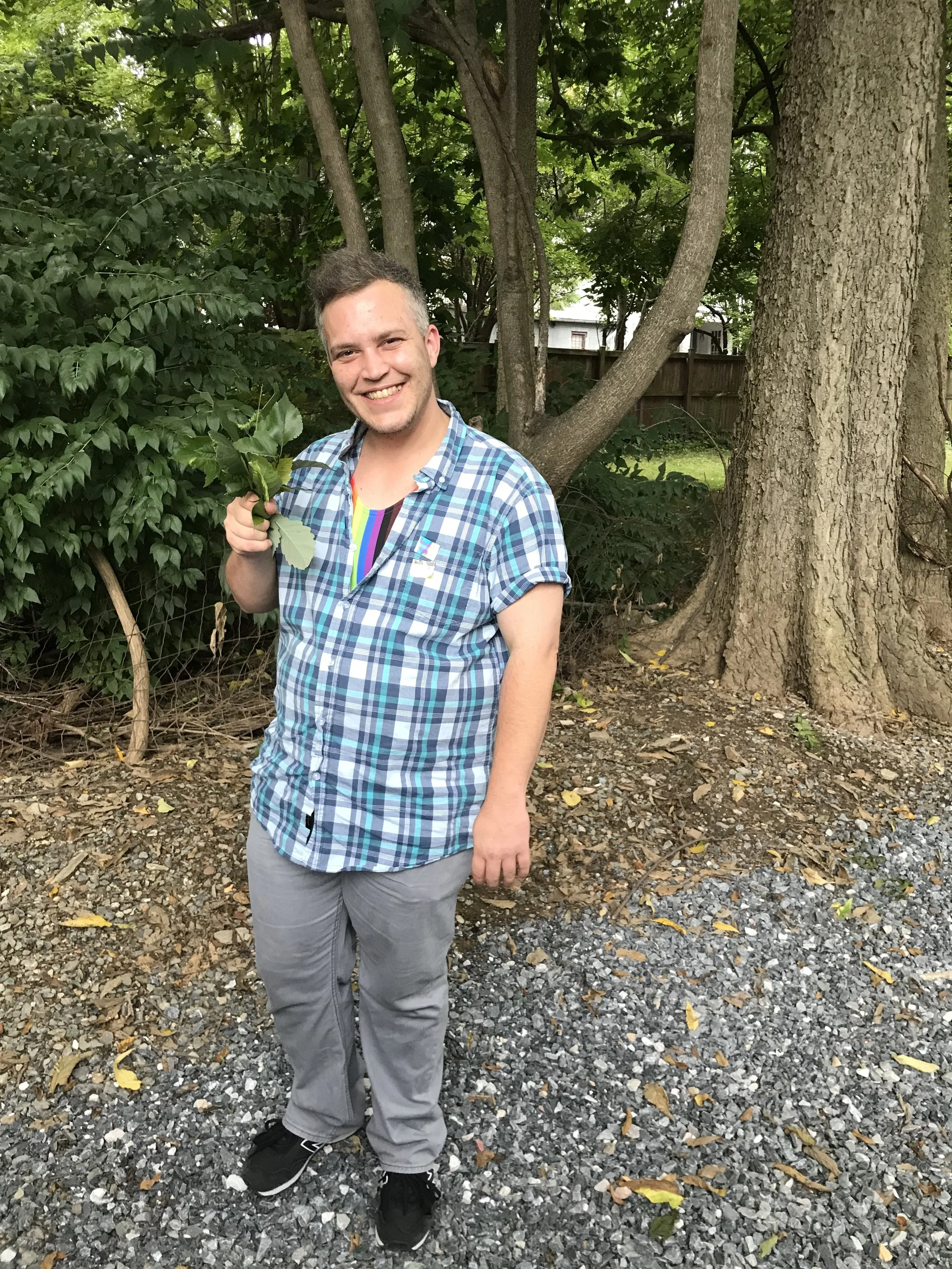 Bastian of AB arts LLC with a bright smile and holding a green leafy bouquet standing outdoors on a gravel surface with trees in the background.