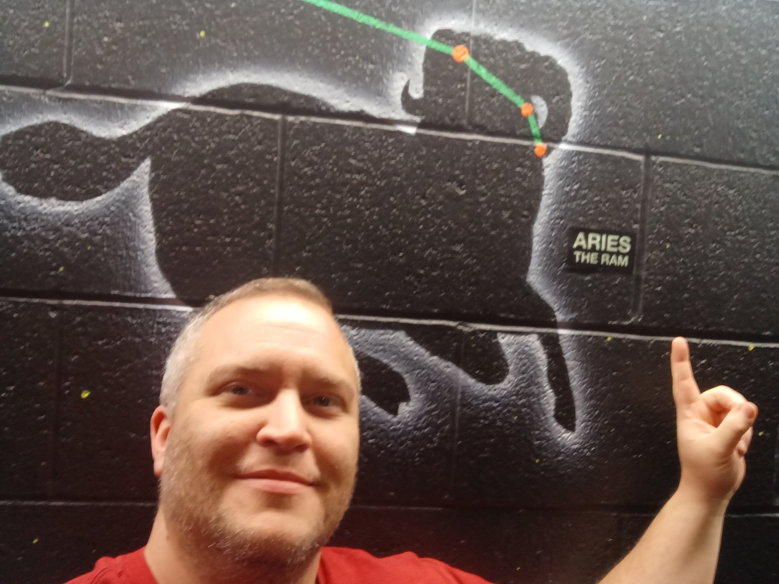 Bastian of AB arts LLC posing with their star sign. They are pointing at a label that reads "ARIES THE RAM."