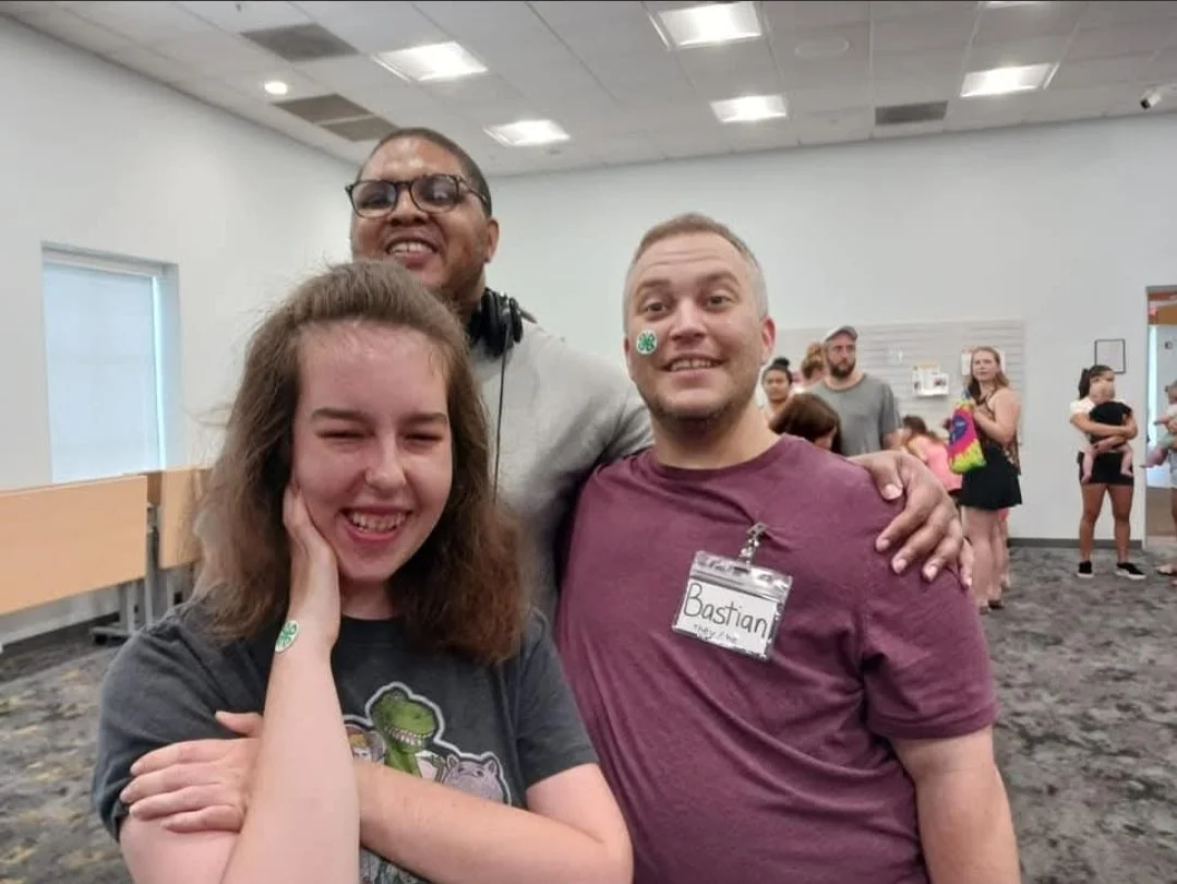 One Black male, one white female and Bastian of AB arts LLC all smiling and posing together for a photo at an indoor Library event.