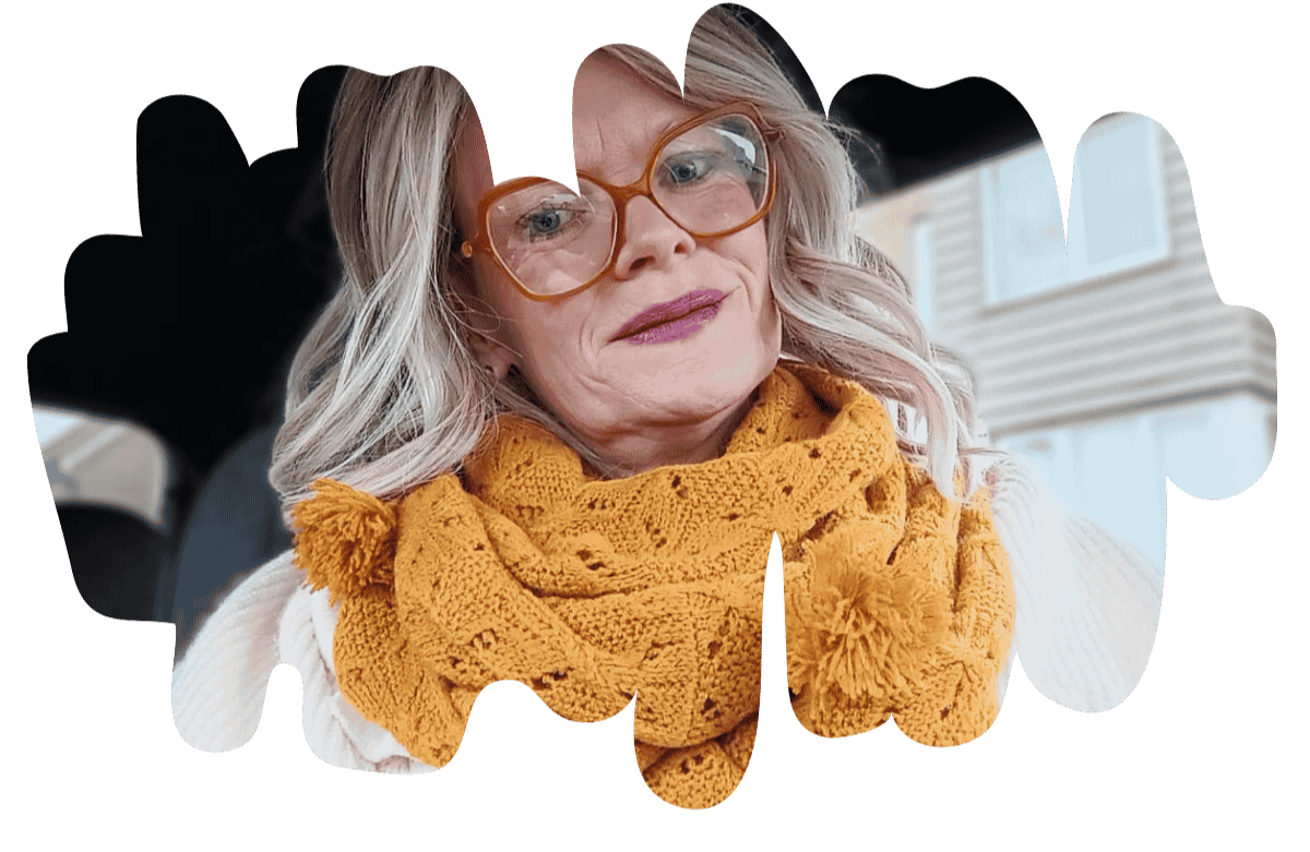 Helena with her blonde wig,  wearing glasses and a mustard-coloured scarf, sitting indoors near a window, showing self-expression and confidence.