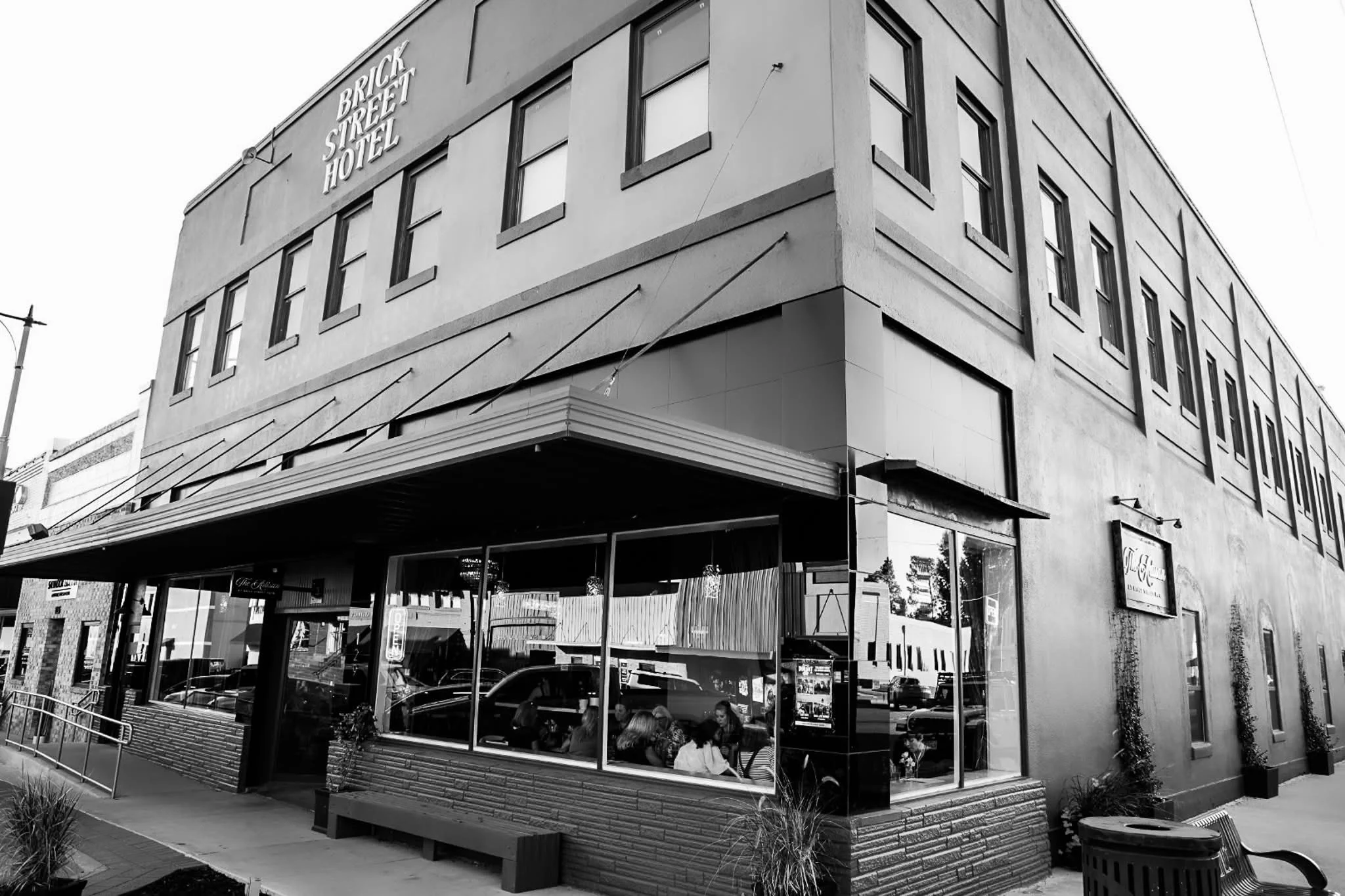 A black and white photo of a corner building with a sign reading 'Brick Street Hotel' on the upper facade. The building has several windows and a reflective glass storefront on the ground level, with patrons sitting inside visible through the windows. There are plants and outdoor seating in front of the building, and a sidewalk with a railing and street furniture. The sky is visible above the building.