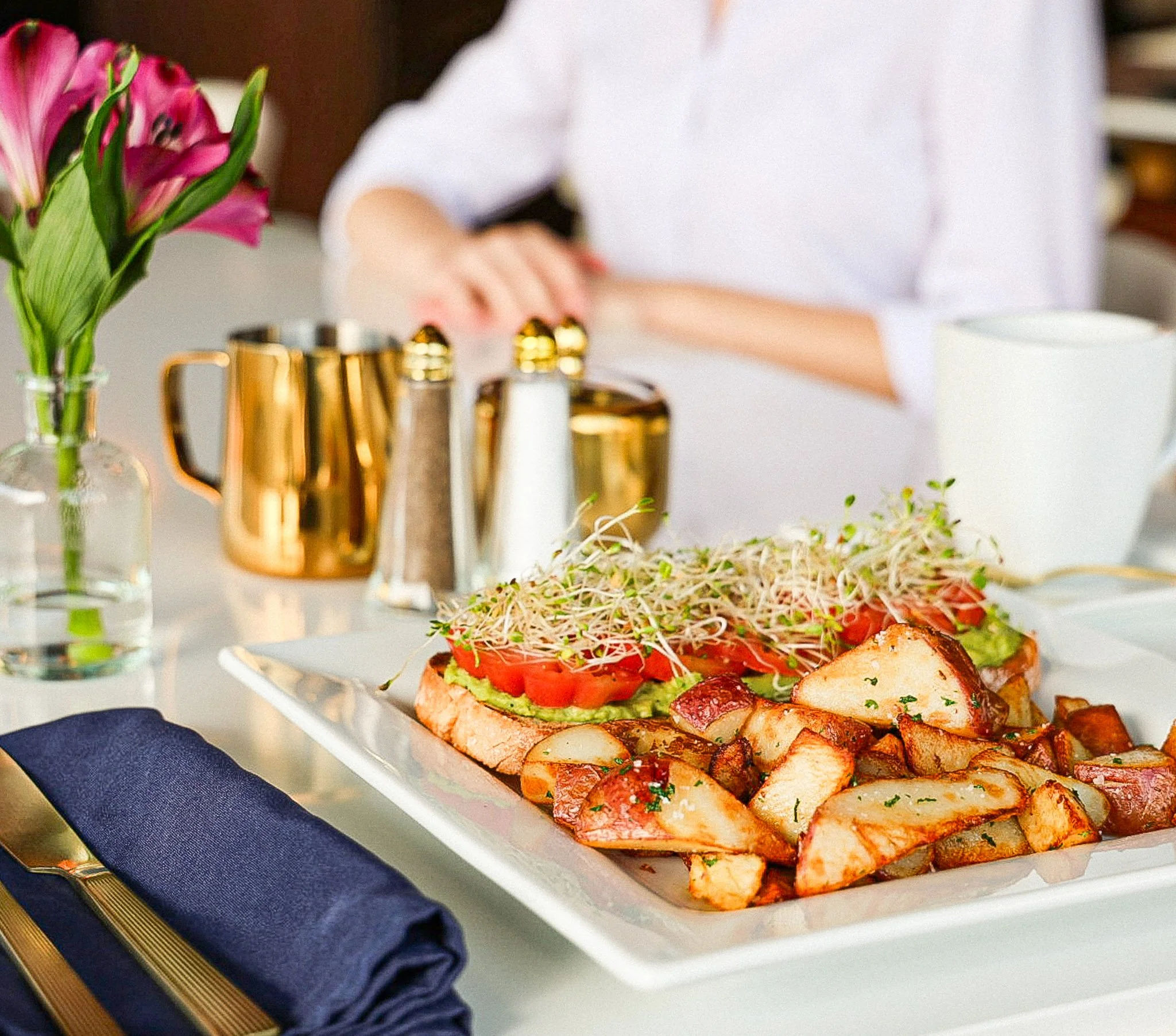 A white rectangular plate with assorted grilled meats and a sandwich with fresh vegetables, placed on a table with a blue napkin and gold-edged plate.