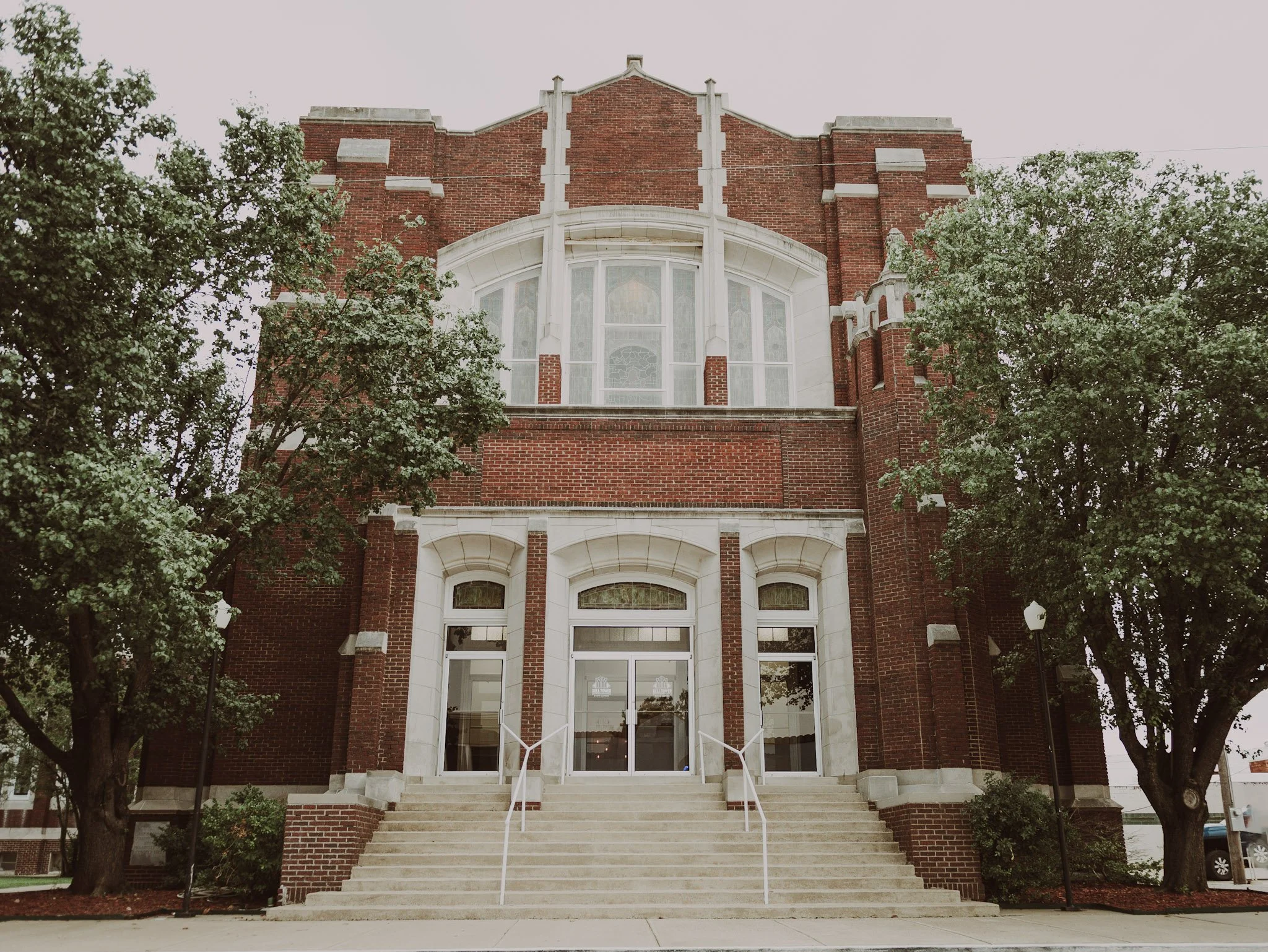 Front view of a historic red brick building with large windows and steps leading to the entrance, flanked by trees.
