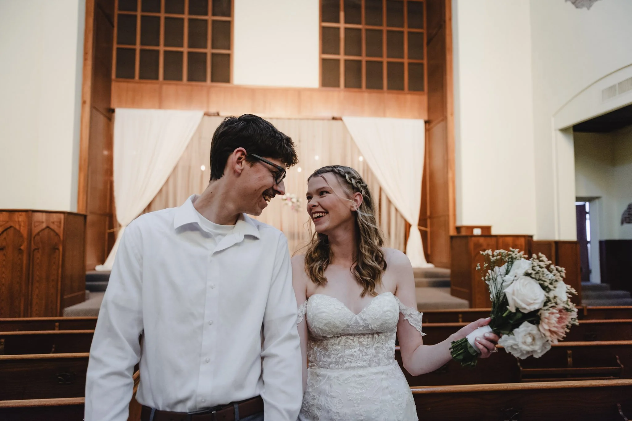 A bride and groom standing together inside a church, smiling and looking at each other. The bride is holding a bouquet of flowers. The background features wooden church pews and an elevated altar area.