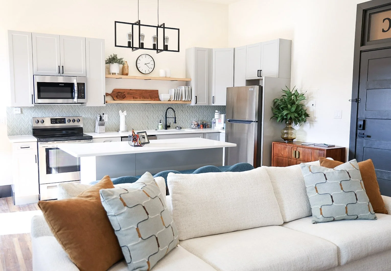 Open-concept kitchen and living room with white sofa, decorative pillows, kitchen cabinets, stainless steel appliances, open shelving, a kitchen island, and a black door.