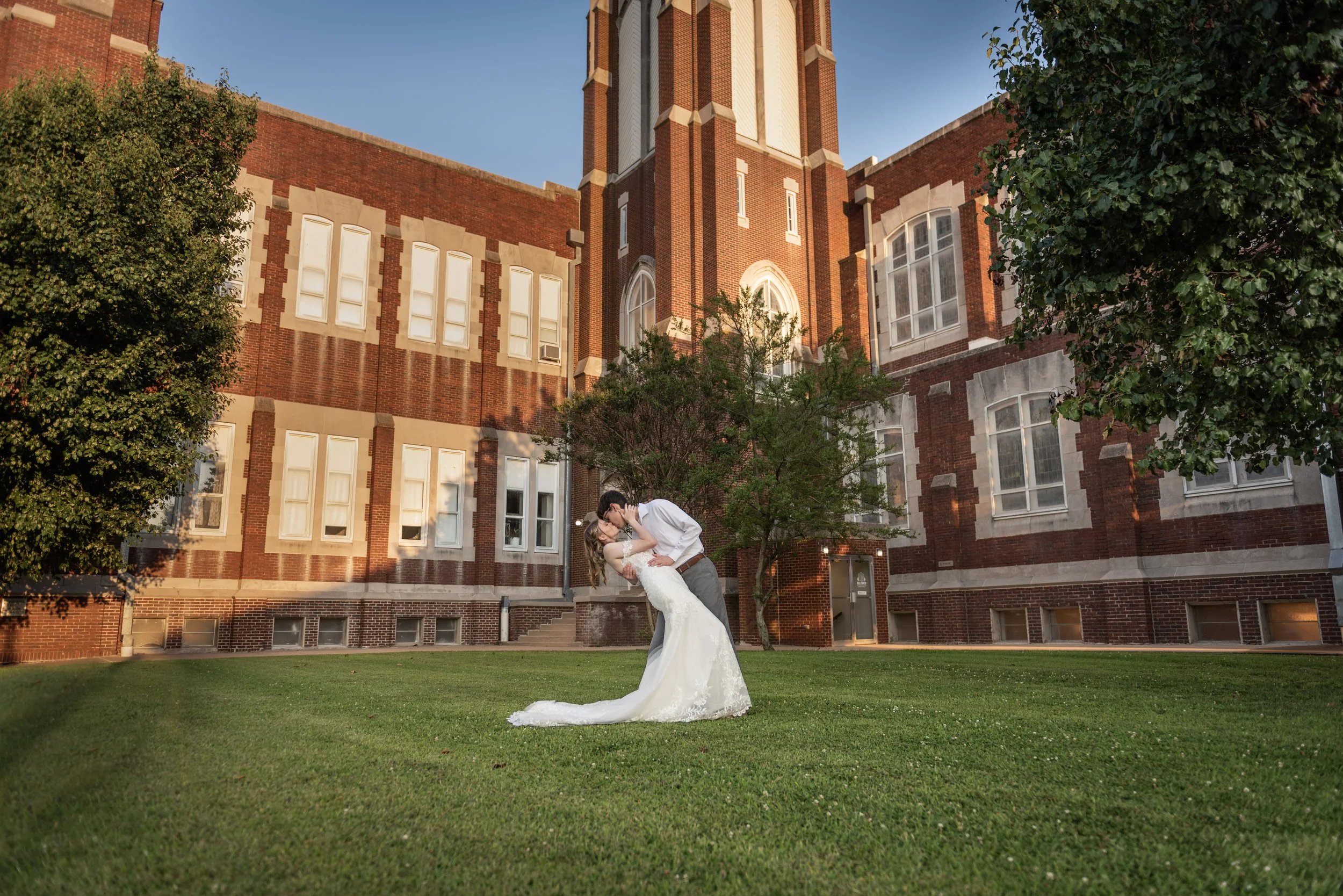 A bride and groom sharing a kiss in front of a red brick building with large windows and trees, during sunset.
