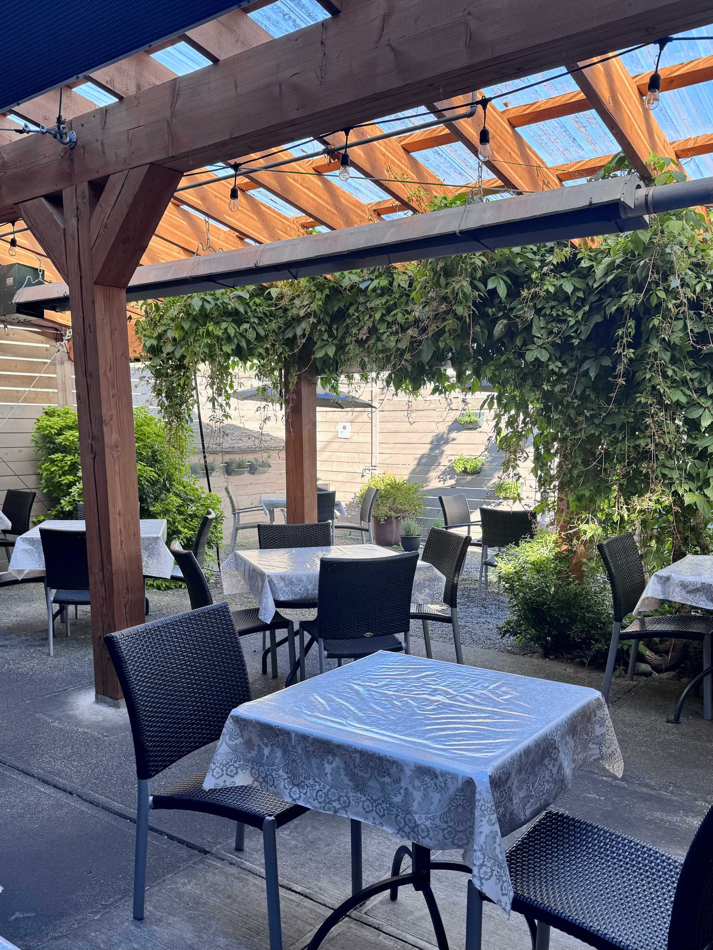 Outdoor patio dining area with tables covered in white patterned tablecloths and black chairs under a wooden pergola with string lights and greenery.