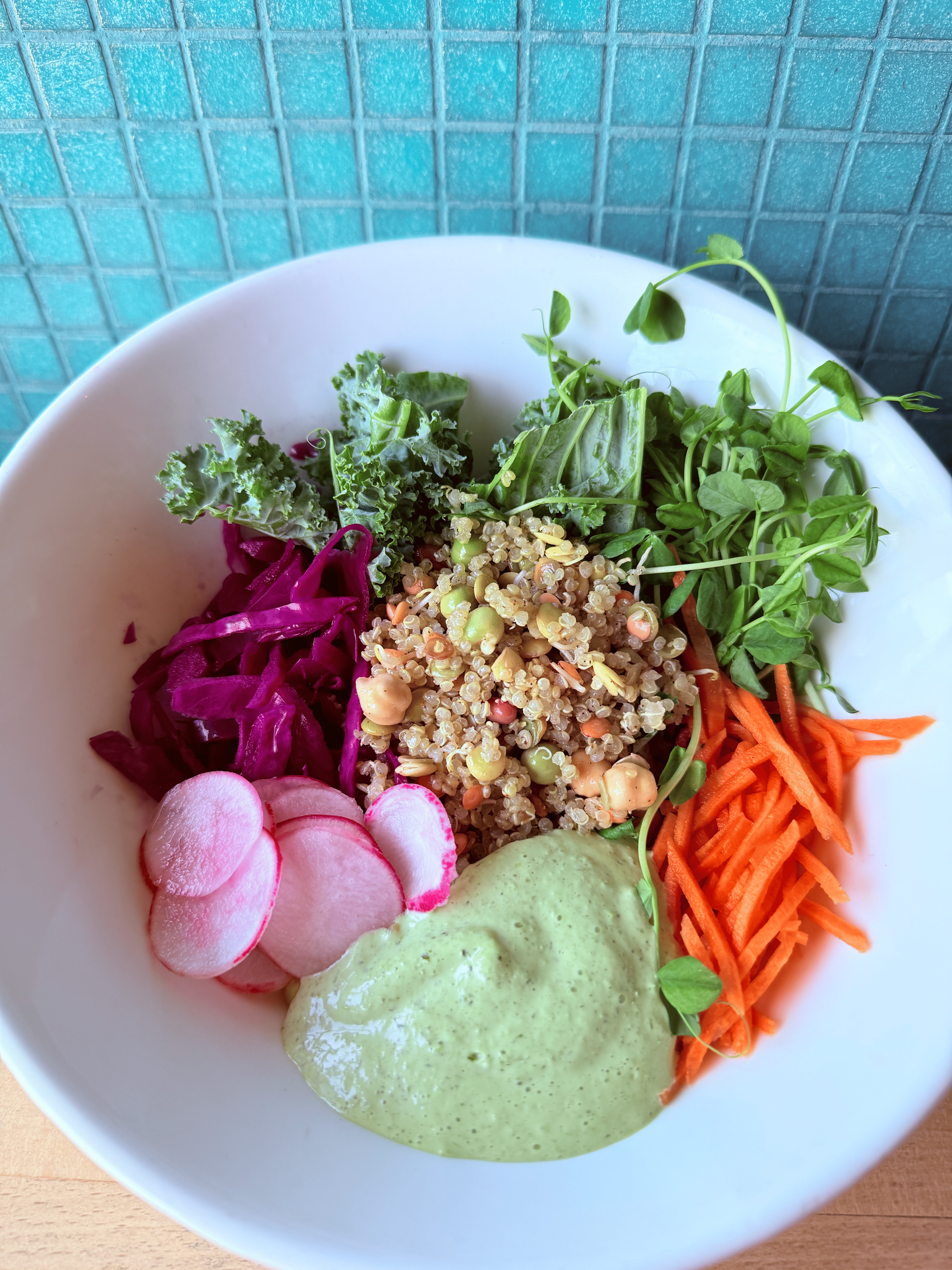 A white bowl containing sliced radishes, shredded purple cabbage, cooked quinoa, chopped carrots, fresh leafy greens, and a dollop of green herb sauce, placed on a light wooden surface.