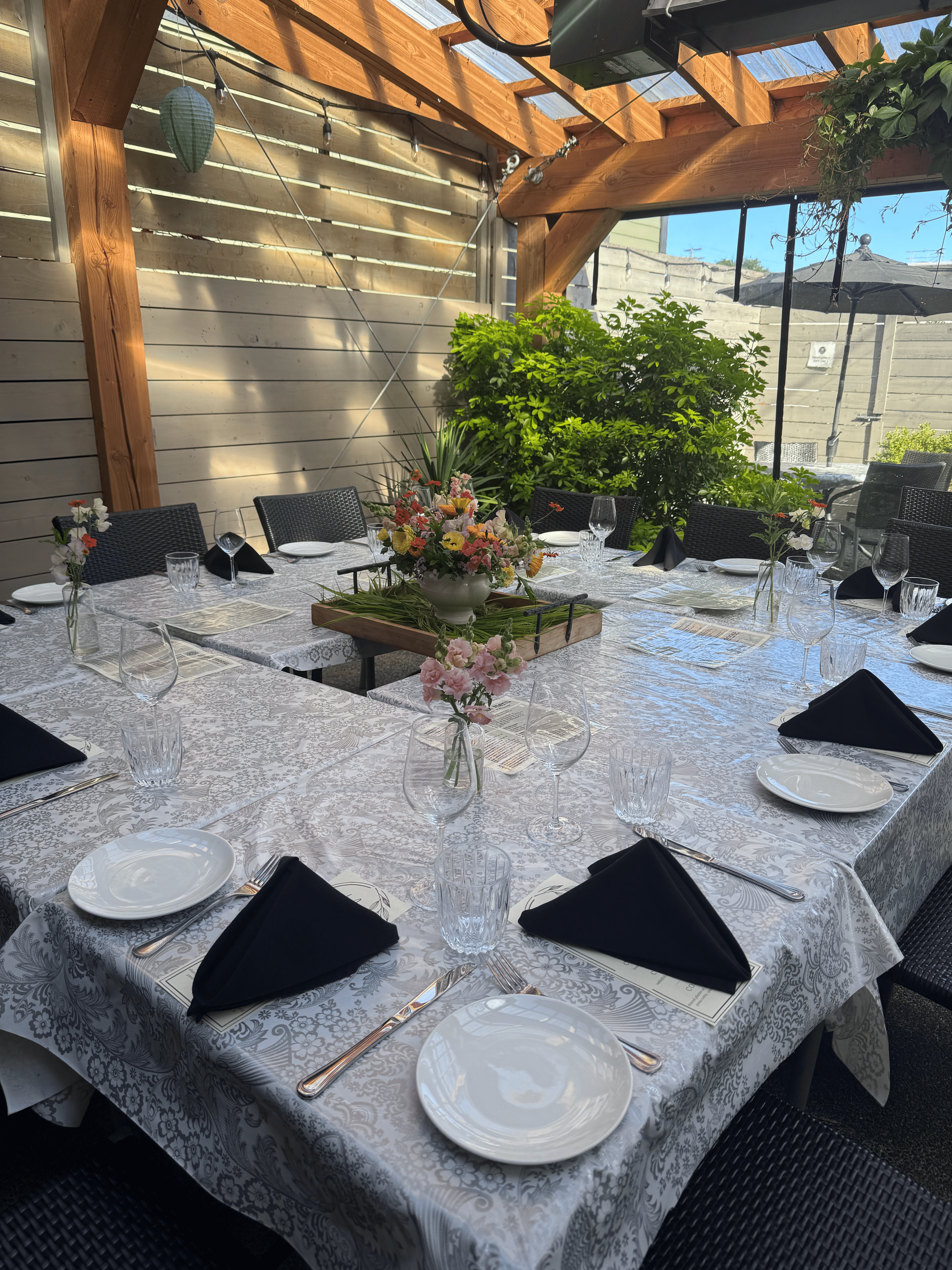 large gather dining table set for a meal on a coutyard patwith white patterned tablecloth, black napkins, clear glassware, white plates, silverware, and a colorful flower centerpiece, in a partially outdoor setting with wooden beams and green plants.