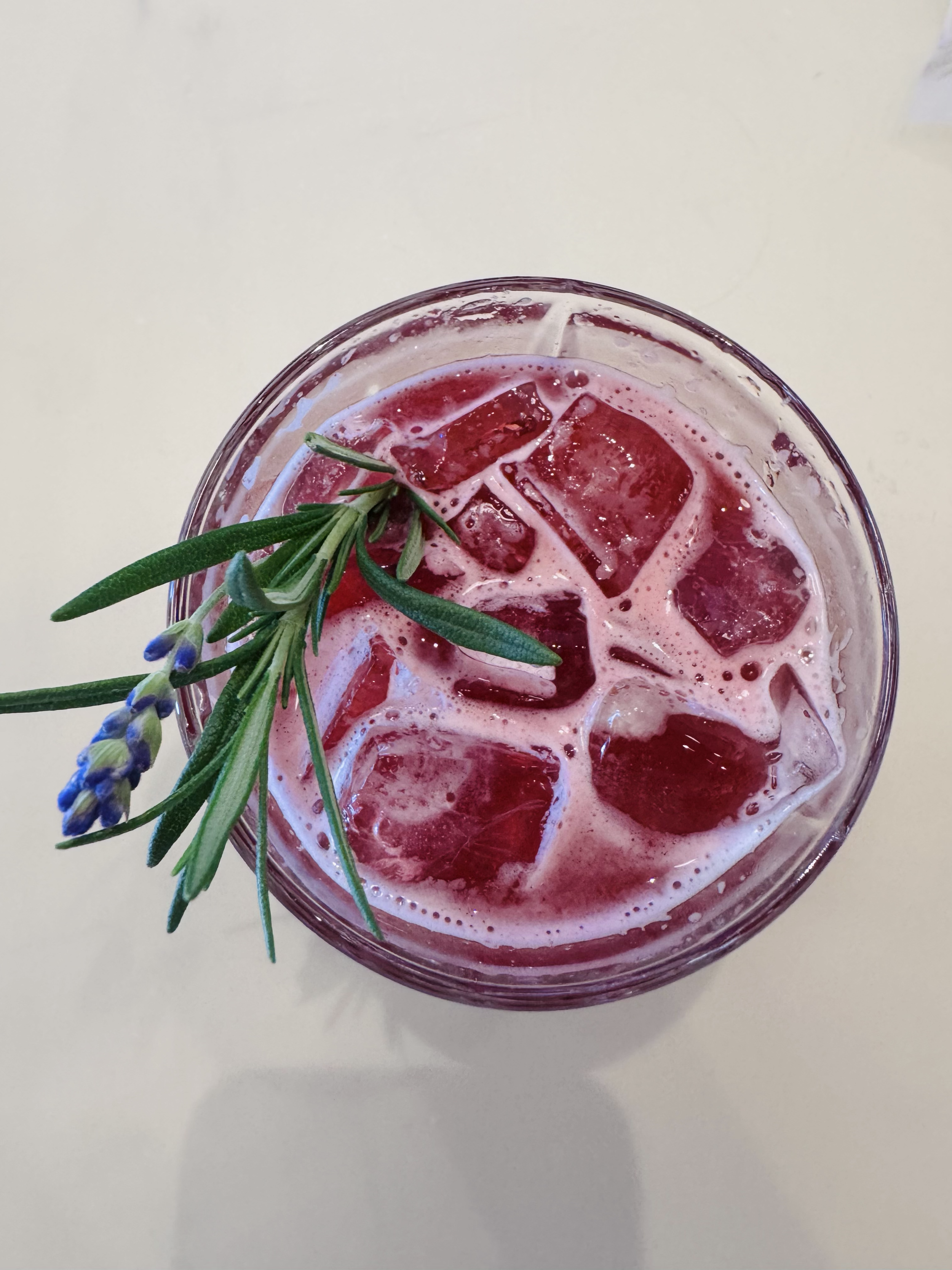 A glass of red beverage with ice cubes, garnished with a sprig of rosemary and a few lavender flowers, on a light-colored surface.