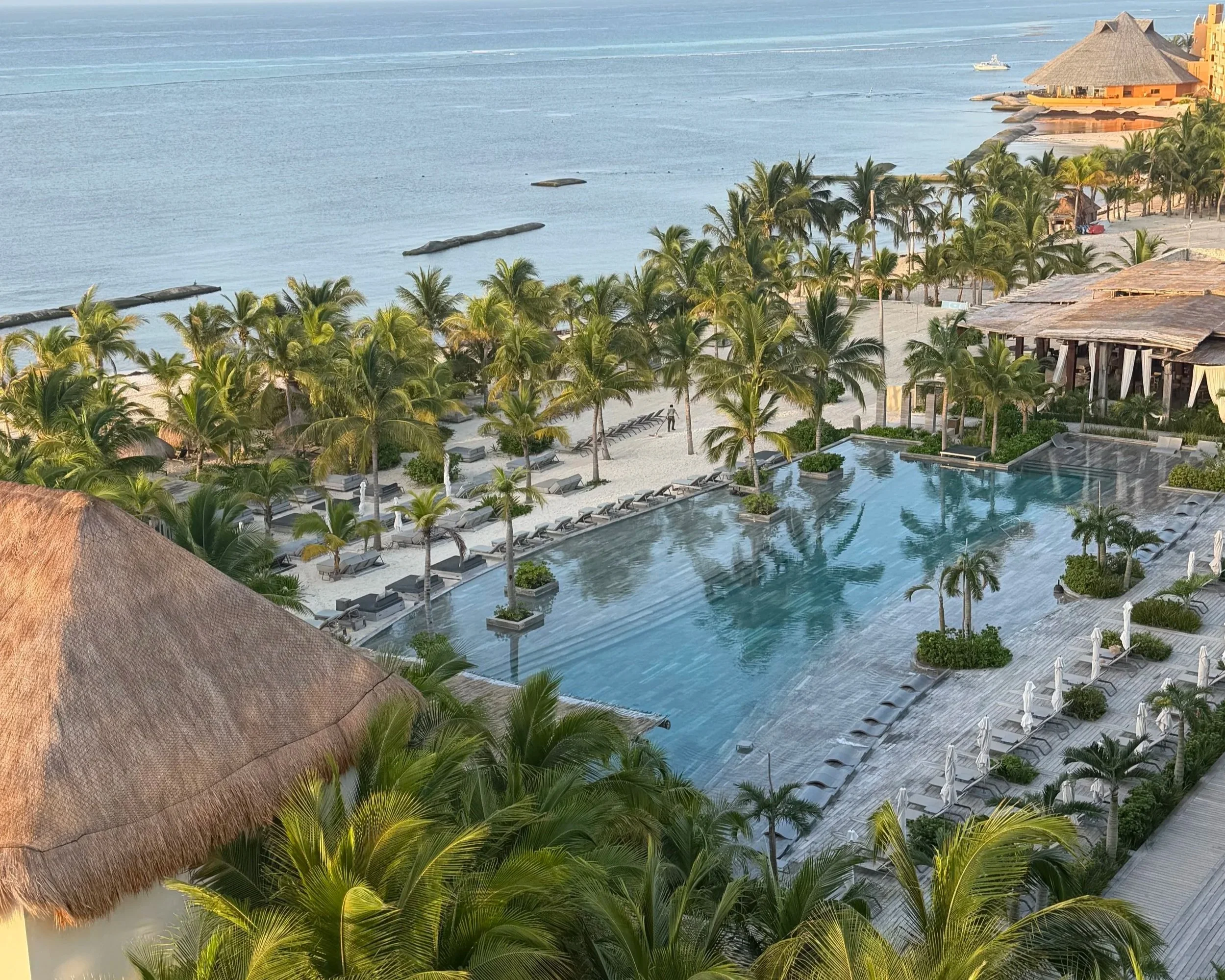 A tropical resort with a large swimming pool surrounded by numerous palm trees, beach chairs, and thatched roof buildings, overlooking the ocean with a boat visible in the water.