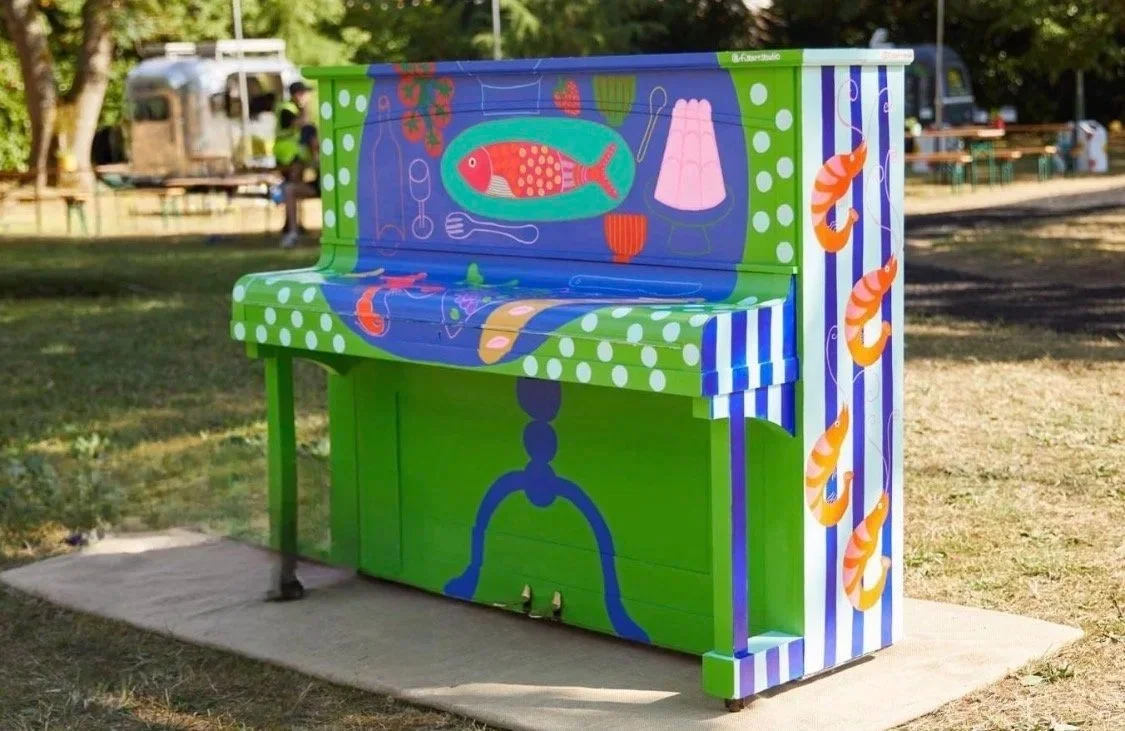 Colorful painted piano outdoors with fish, shrimp, and food motifs in a park setting.