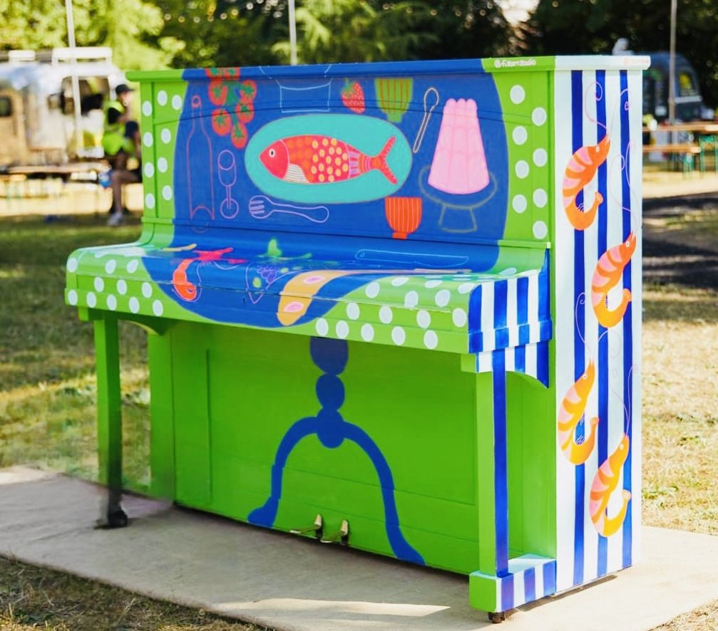 Colorful painted piano decorated with fish, shrimp, and seafood-themed illustrations, outdoors in a park.