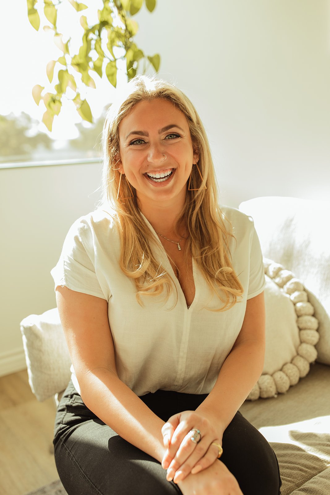 A woman with blonde hair sitting on a bed, smiling, with sunlight coming through a window behind her.