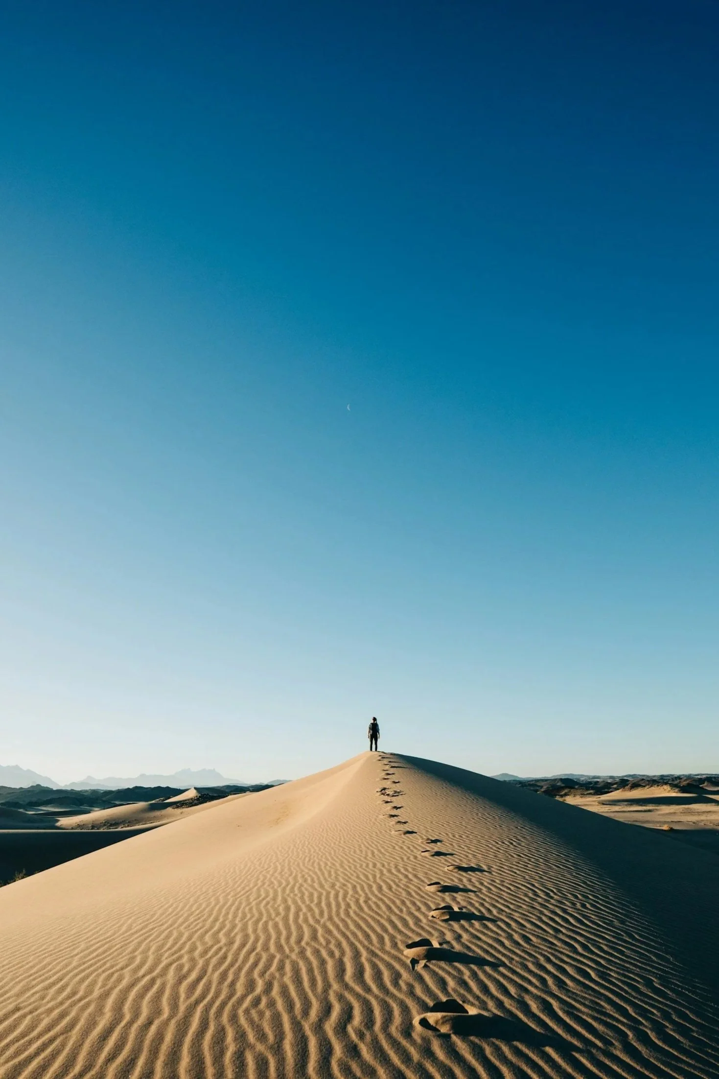 A person with a backpack walking on a sand dune in a desert, leaving footprints behind, under a bright blue sky with a thin crescent moon visible.