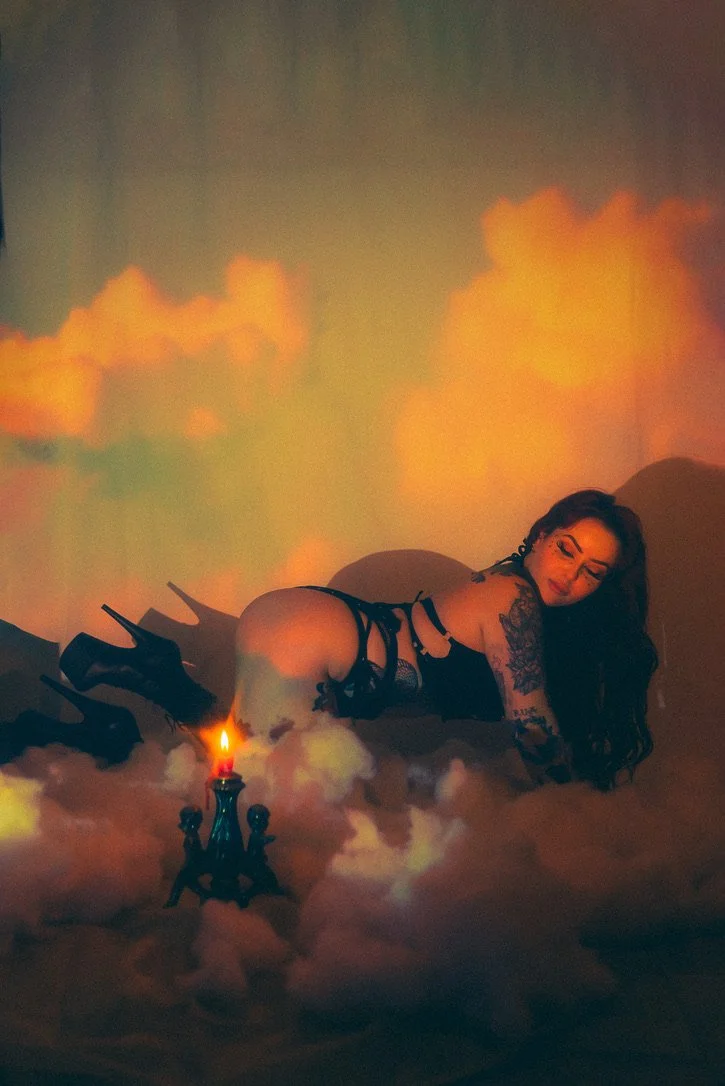 A woman with tattoos and dark hair wearing black lingerie and high heels, posing on all fours among clouds or fog with a lit candle nearby, with orange clouds projected on the background.