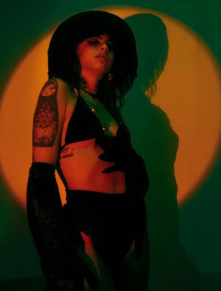 A woman with dark hair and tattoos, wearing black gloves and lingerie, posing against a dark background with a yellowish circular light and green shadow, in a provocative stance.