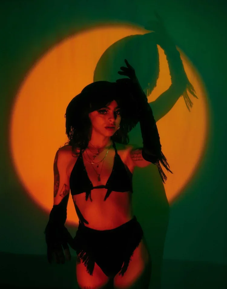 A woman dressed in black lingerie with tattoos, standing in front of a circular yellow-orange light projection, creating a silhouette shadow on the wall behind her.