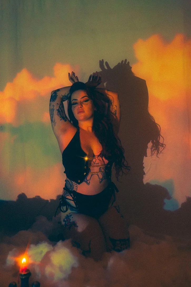 A woman with long curly hair and tattoos on her arms and legs is posing in lingerie with her eyes closed, against a backdrop of orange clouds projected on the wall. There is a lit candle and fog at her feet.
