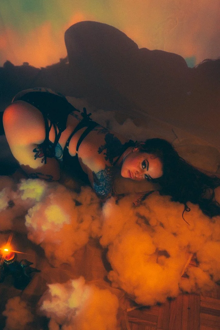 A woman with dark curly hair and tattoos on her arms and leg, dressed in black lingerie, lying on the floor amidst orange smoke. She is looking at the camera with an intense expression under atmospheric lighting.