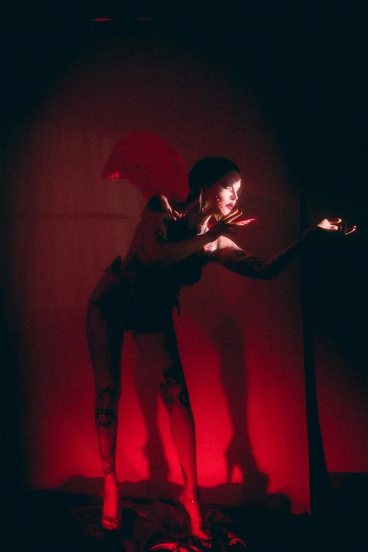 A woman dressed in dark clothing with tattoos on her legs, illuminated by red light, posing with one arm extended.