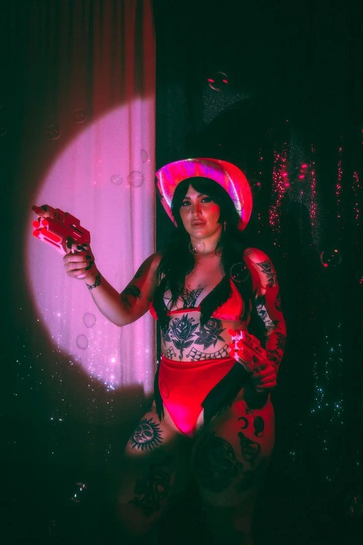 A woman with dark hair and tattoos wearing a red bikini, a pink cowboy hat, and holding a gun, standing in front of a dark background with pink and red lighting.