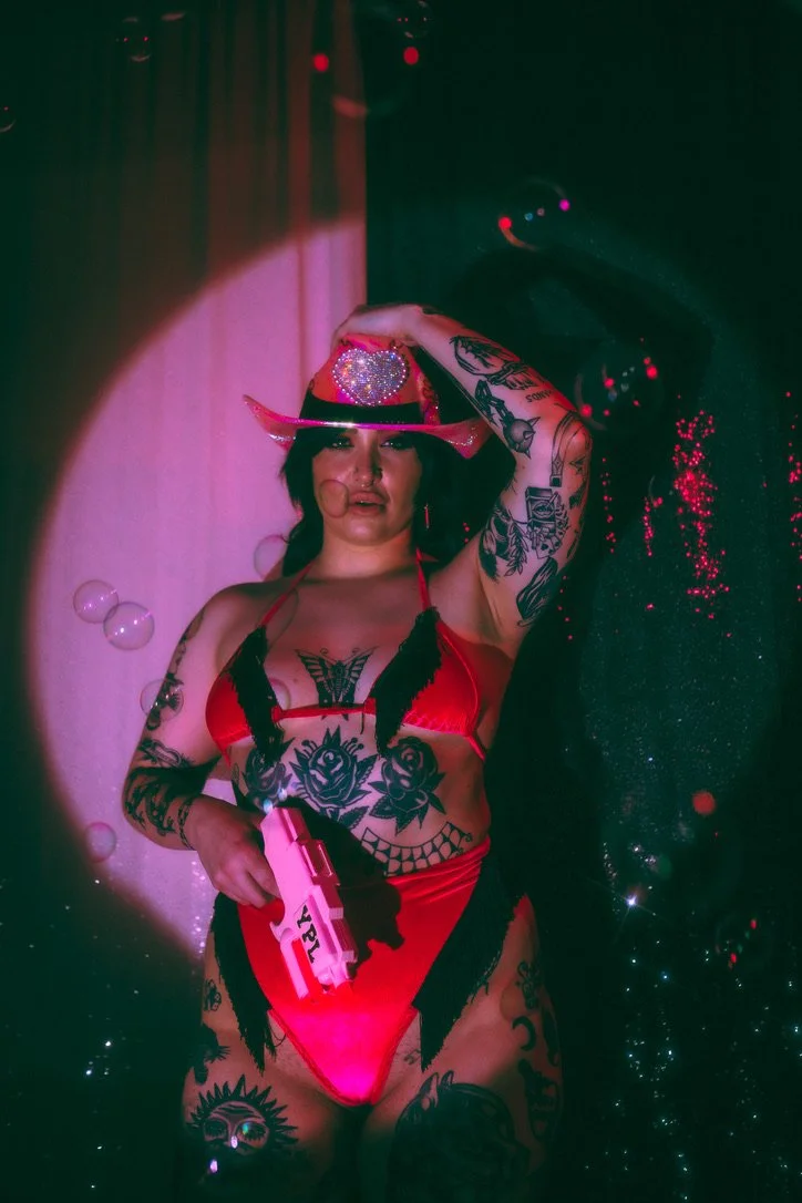 A woman wearing a red swimsuit, black feathered accessories, and a pink cowboy hat with rhinestones, holding a pink gun with the word 'YPL' on it, surrounded by colorful lighting and bubbles.