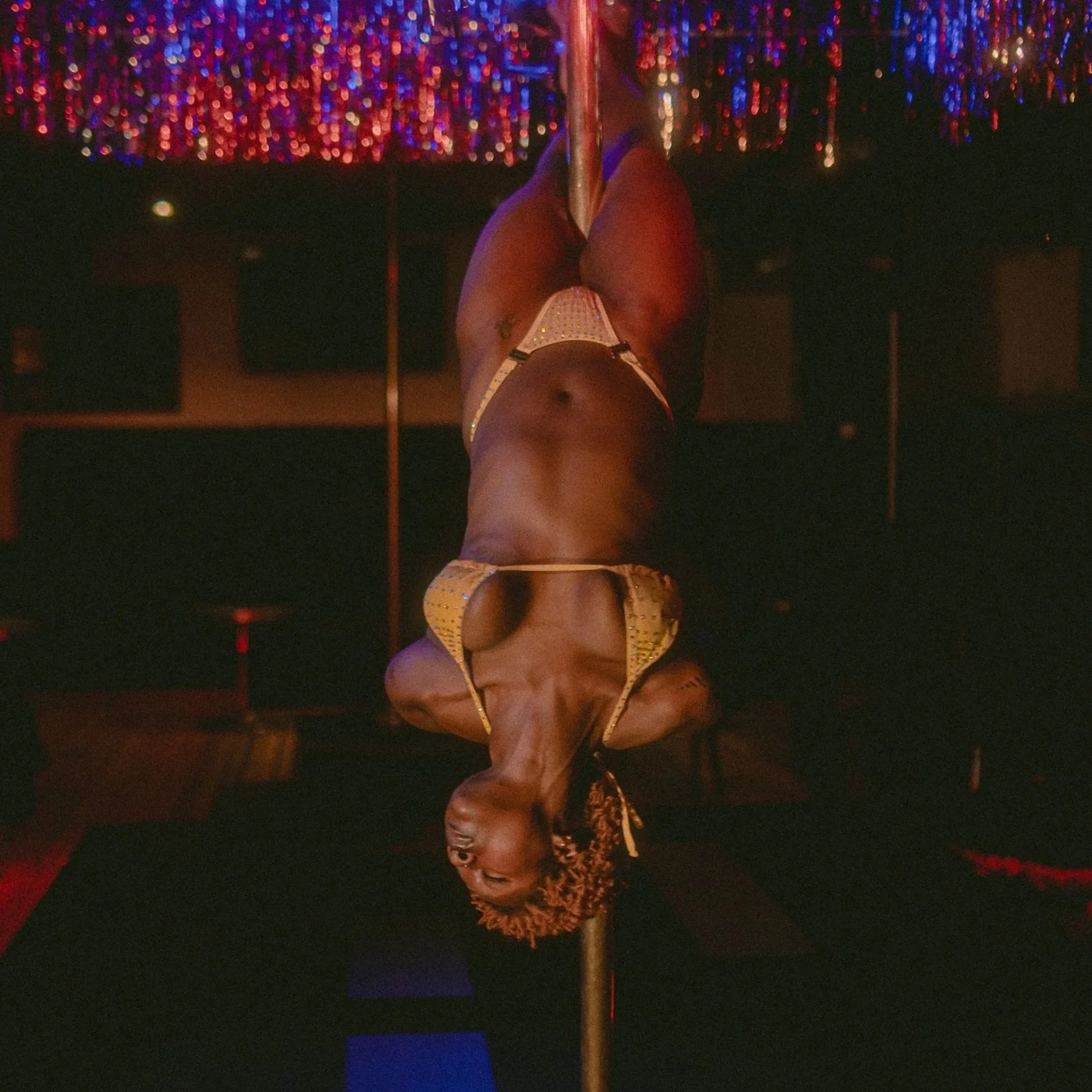 A person with curly hair, wearing a shiny yellow outfit, performing a pole dance on a stage with colorful, glittering lights in the background.