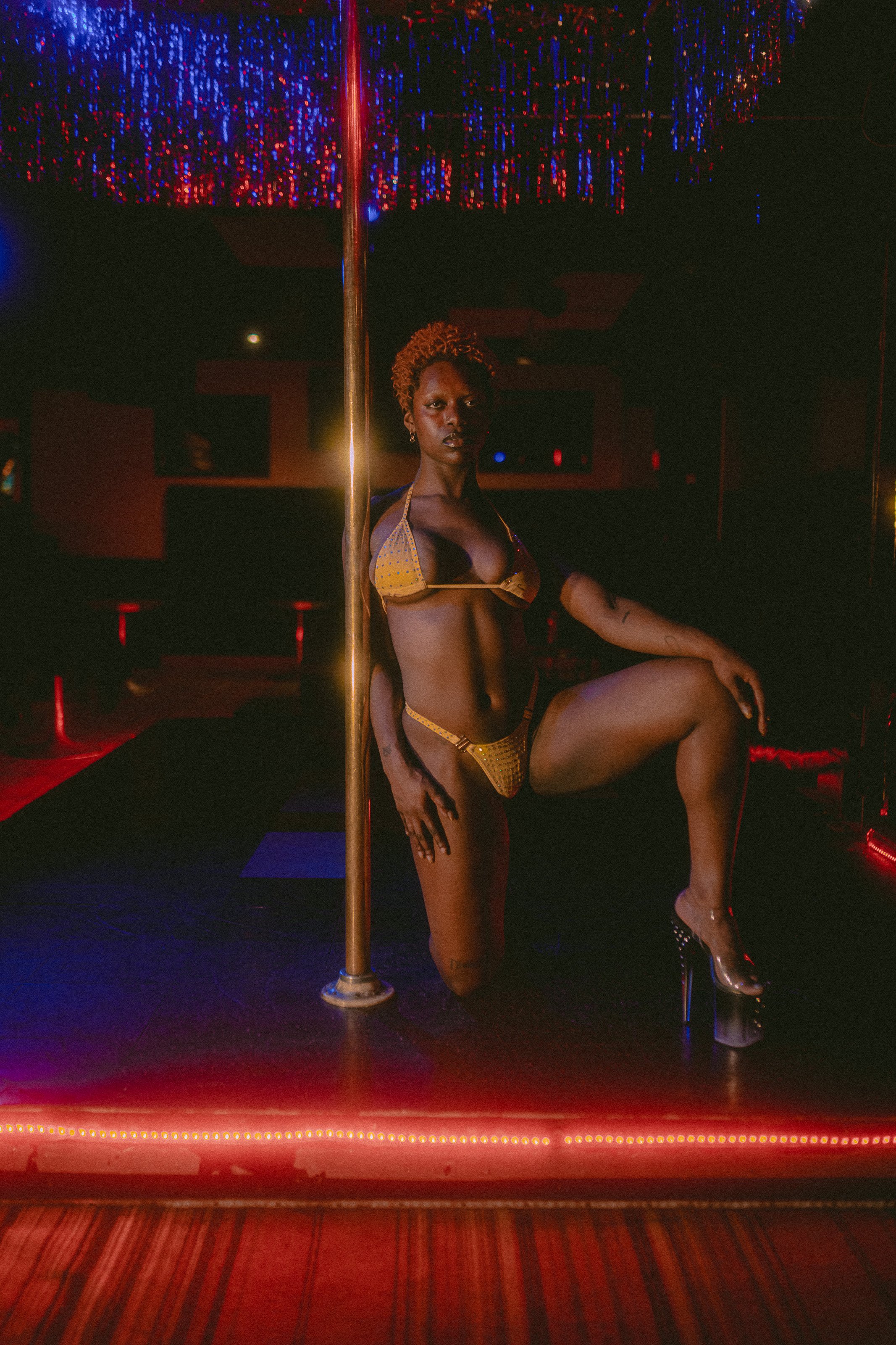 A woman with curly hair and tattoos sitting on a stool in a dimly lit room with a pole, wearing lingerie and high heels, illuminated by red and purple lights.
