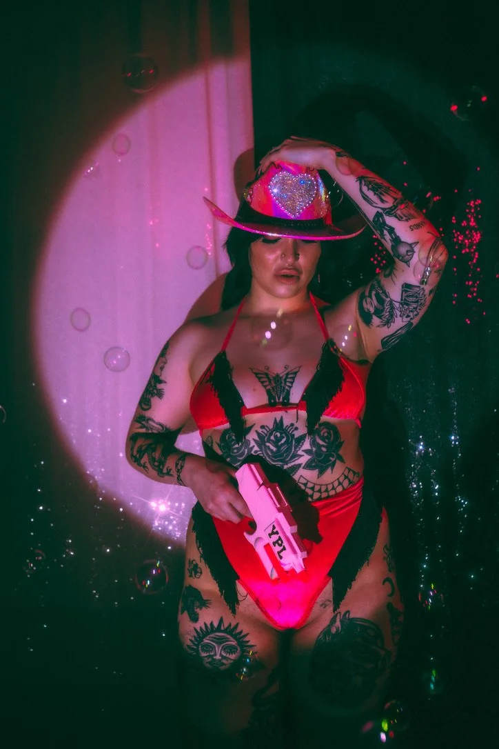 A woman dressed in red and black lingerie with tattoos, wearing a hat with a heart-shaped decoration, holding a toy gun labeled 'YPL', in a colorful, sparkly setting.