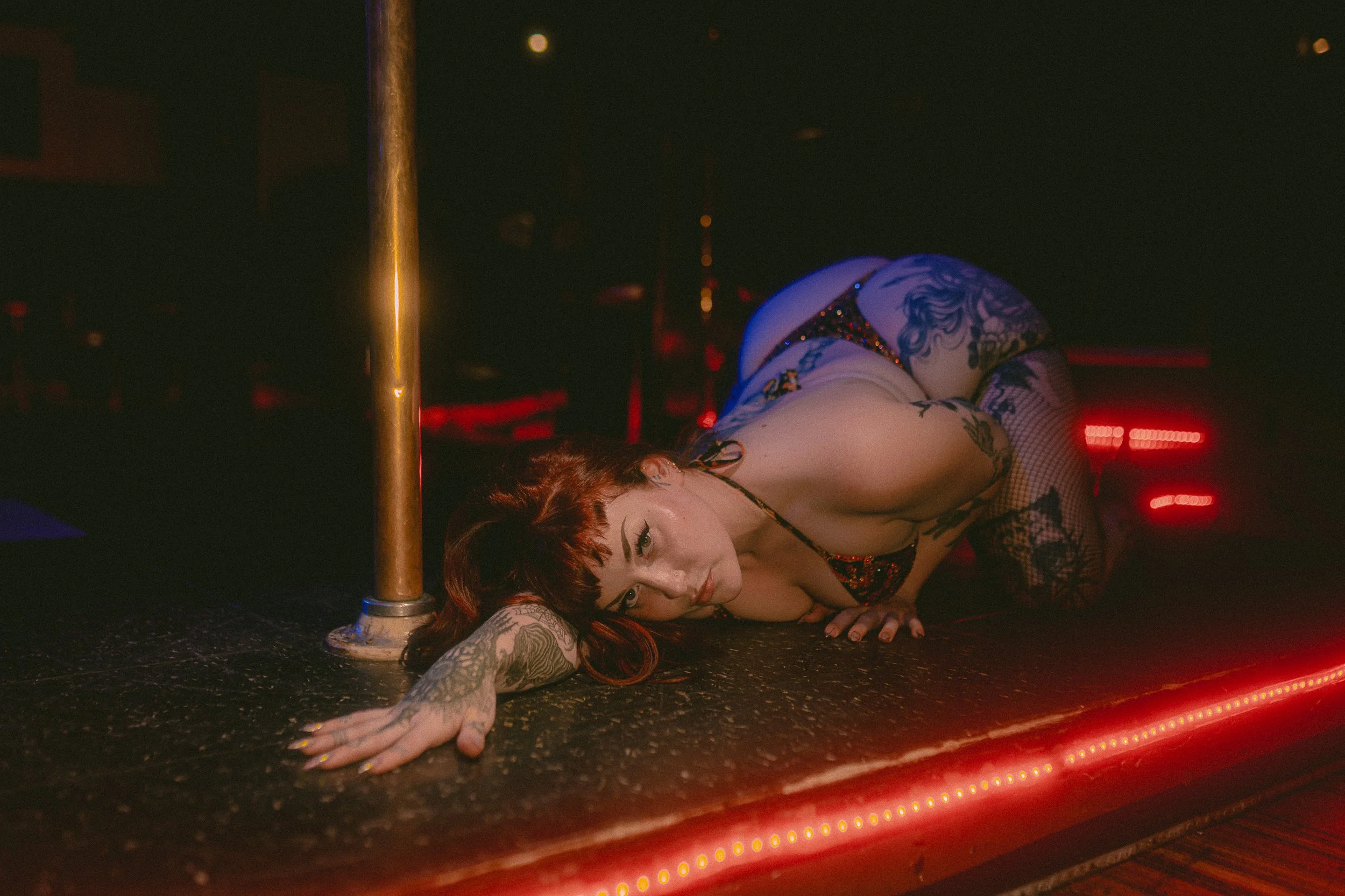 A woman with tattoos and red hair lies on the floor of a dimly lit bar or club with a pole nearby. She is wearing fishnet stockings and a revealing outfit, looking directly at the camera with a relaxed expression.