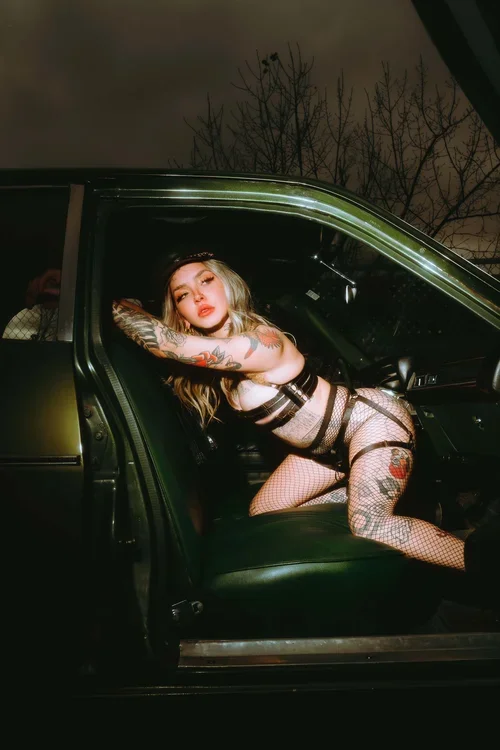 A woman with tattoos and fishnet stockings posing inside a dark green car at night.