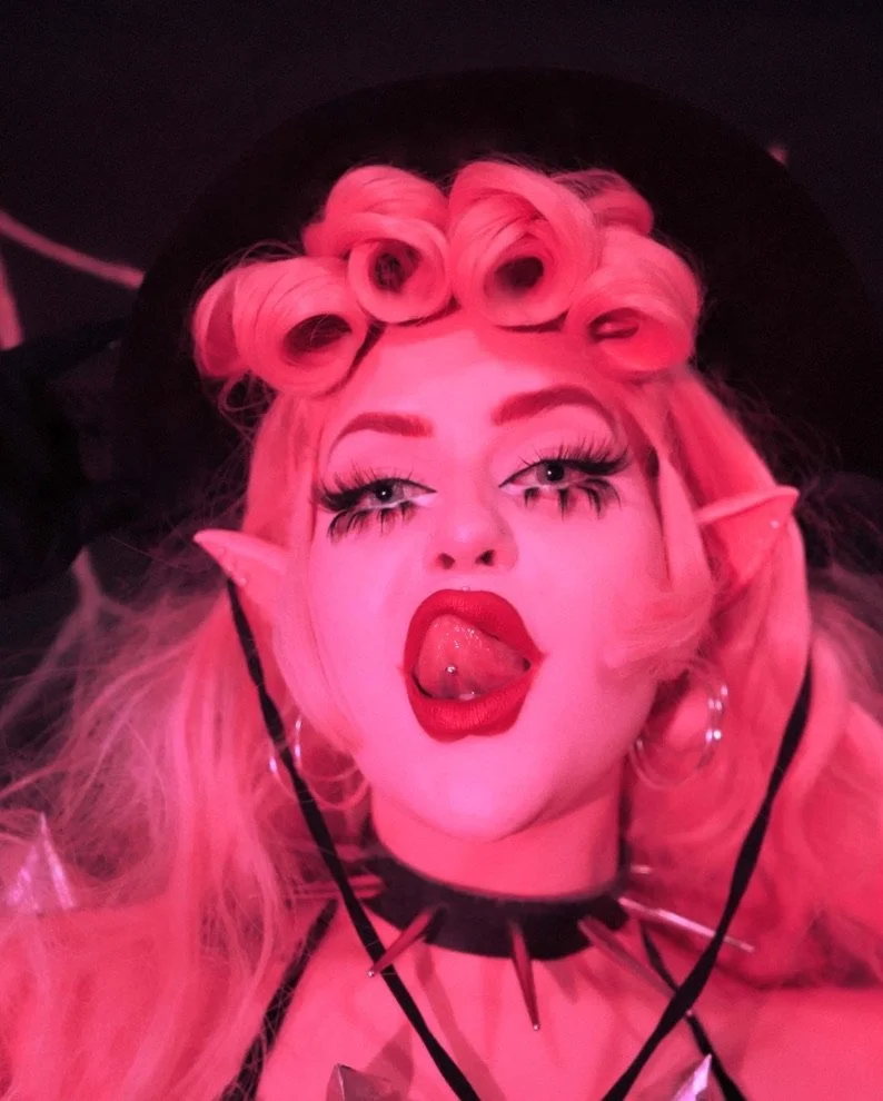 A person with pink hair styled in curls, elf-like pointed ears, and wearing bold makeup, sticking out their tongue with a piercing, in a dark setting with pink lighting.