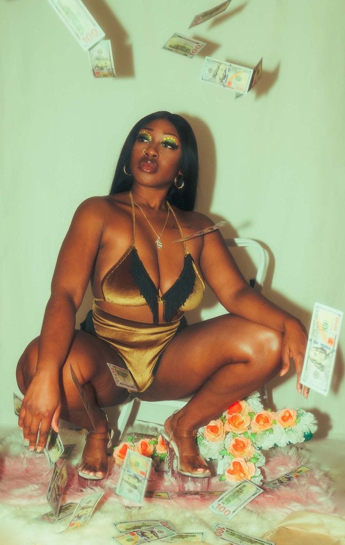 A woman is squatting in front of a white wall with falling money around her, wearing gold and black lingerie, clear high heels, and accessories, with a basket of flowers beneath her.