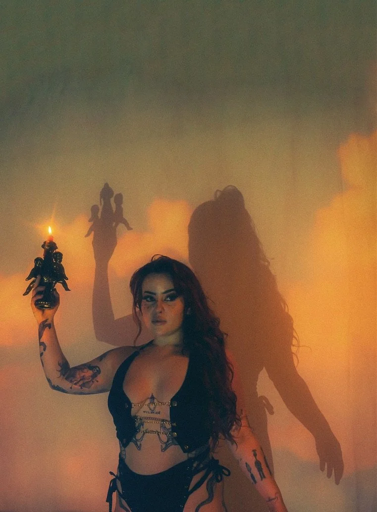 A woman with dark hair and tattoos holding a short-trumbed torch in front of a wall with colorful lighting. Her shadow is cast on the wall behind her.