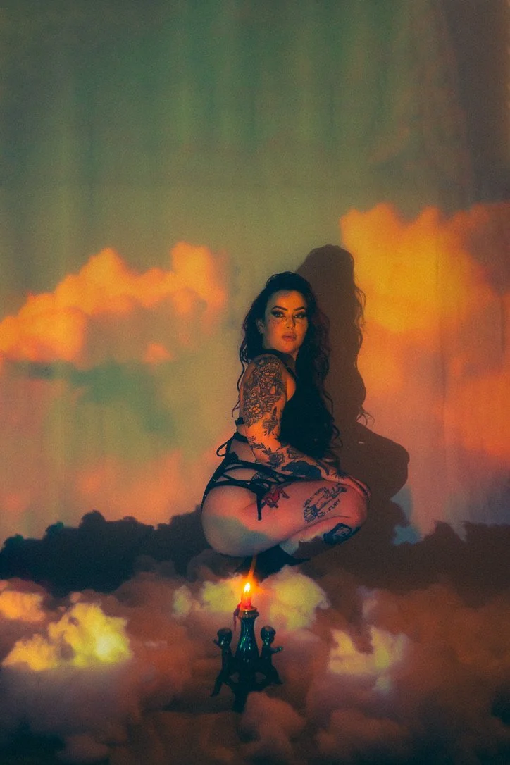 A woman with tattoos sitting on clouds with a chandelier and a candle in front of her. A colorful, cloudy sky background is projected behind her.