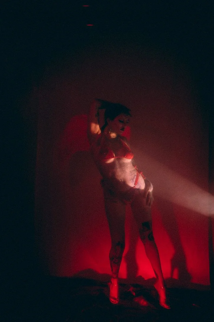A woman in red lingerie and high heels poses under red lighting in a dark room.