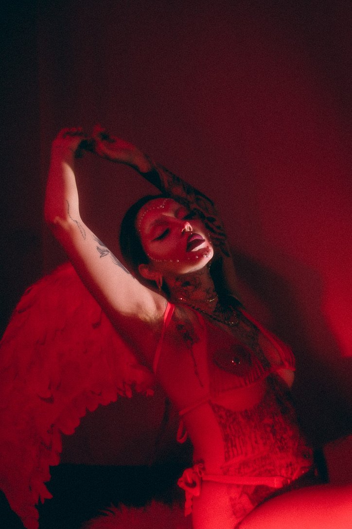 A woman dressed in red with angel wings, dramatic makeup, and tattoos, posing with her arm raised against a dark background lit with red lighting.