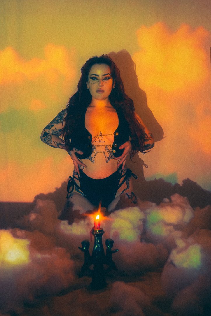 A woman with tattoos on her arms and chest, standing in a cloud-like setting with a dark candelabra and a lit candle in front of her. The background has a warm, orange and yellow glow, creating a mystical or surreal atmosphere.