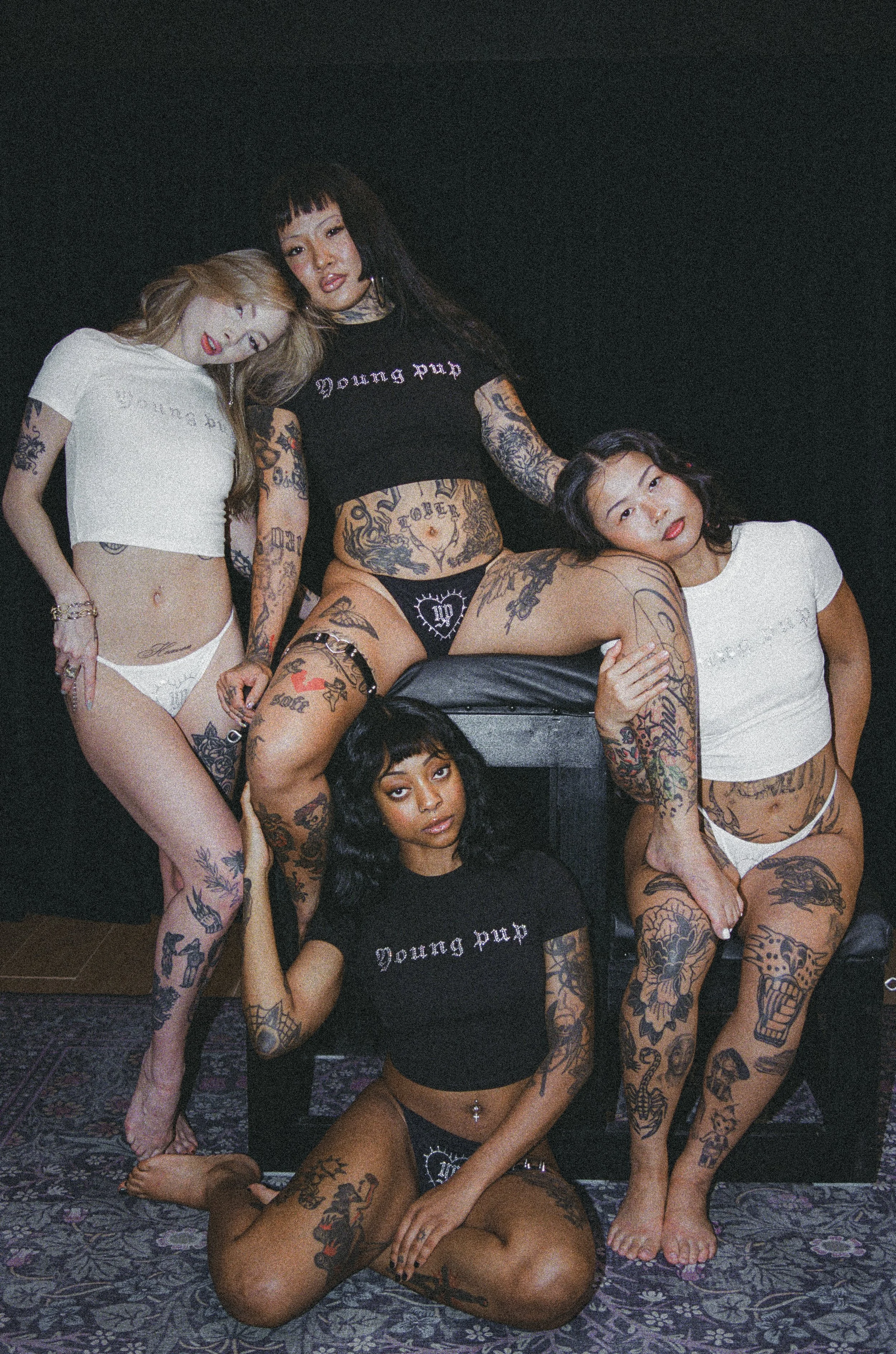 Group of four heavily tattooed women posing together on a patterned carpet in a dimly lit room.