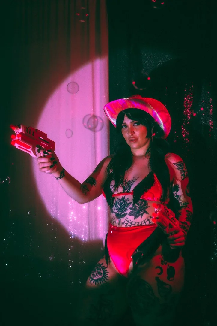 A woman with dark hair and tattoos, wearing a red cowboy hat and red lingerie, holding two toy guns and aiming them. She is standing in front of a backdrop with pink and black lighting effects.